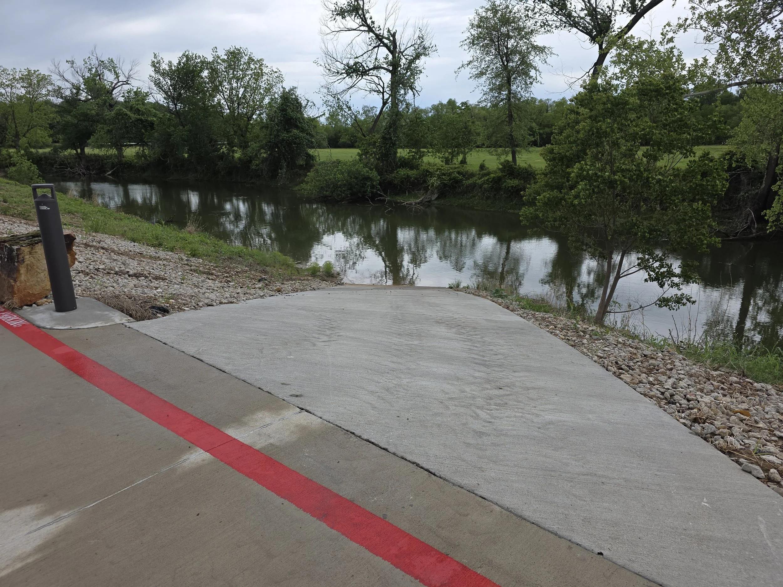 A scenic view of a river with lush green trees on both sides, a cloudy sky above, and a walking path with a red line on the edge in the foreground.
