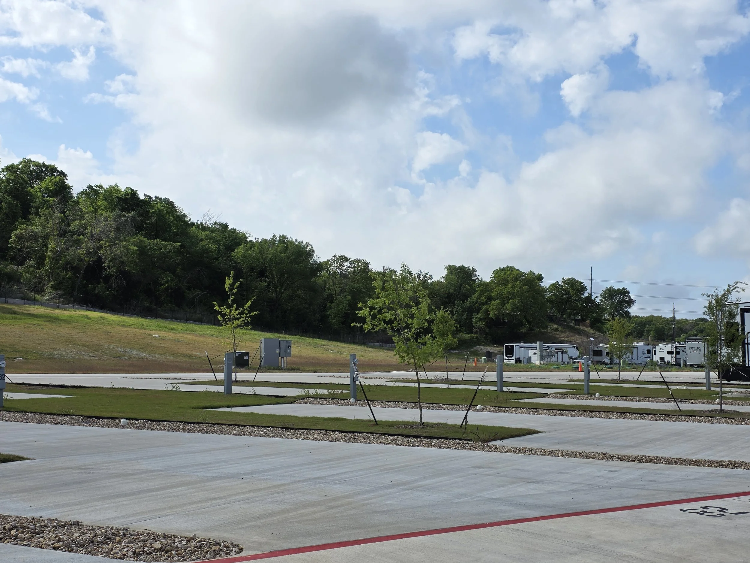 A parking lot with a row of RVs and trailers parked near a hillside with green trees. The sky is partly cloudy with large white and gray clouds.