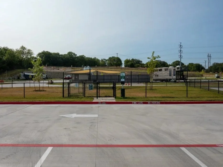View of a fenced RV resort dog park with a recreational vehicle, a small building, and trees in the background. The foreground shows an empty parking lot with red-painted curbs and a fire lane.