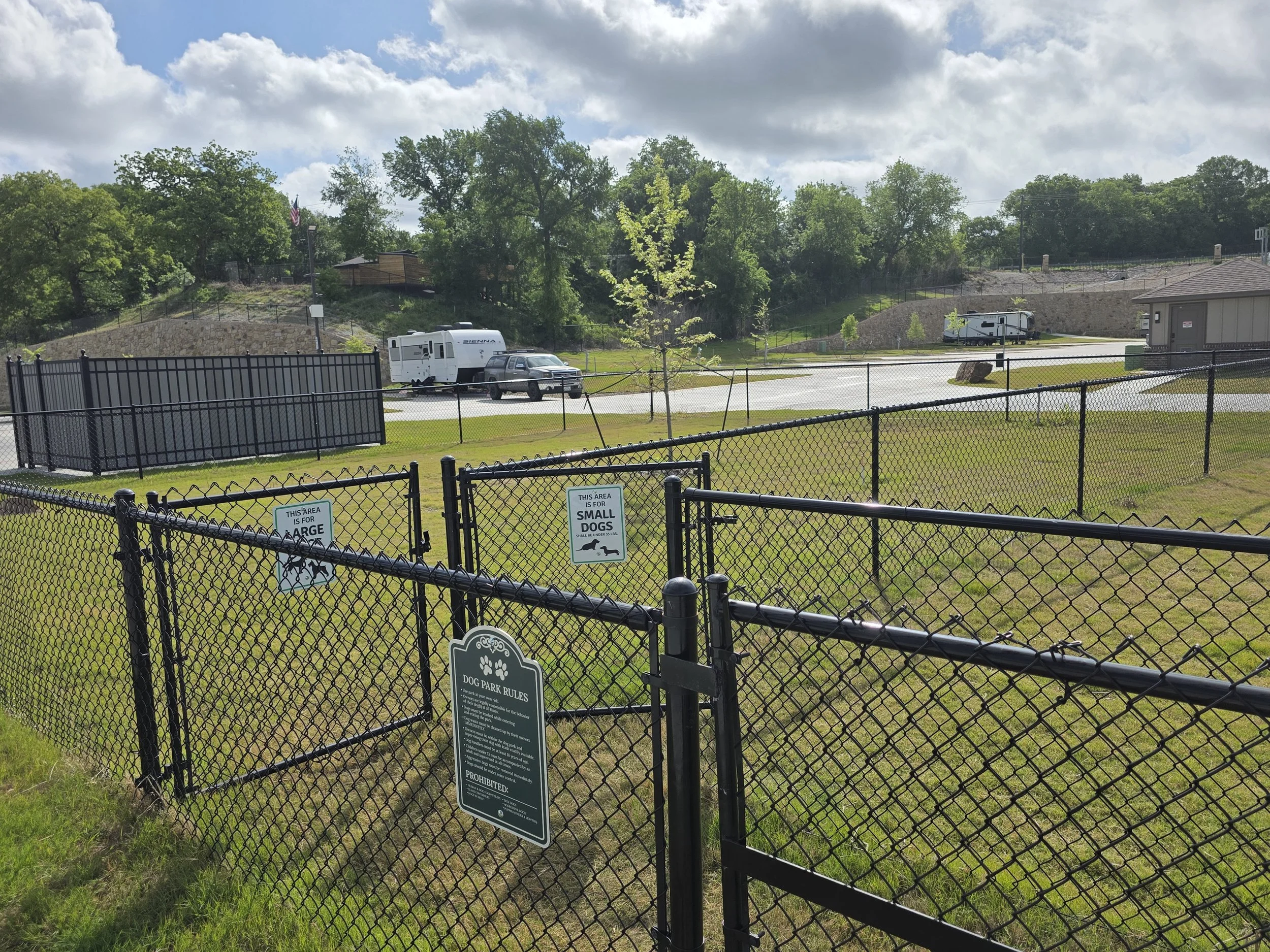 Entrance gates of dog park showing signs with rules and signs for small and large dog enclosures, RVs visible in the background.