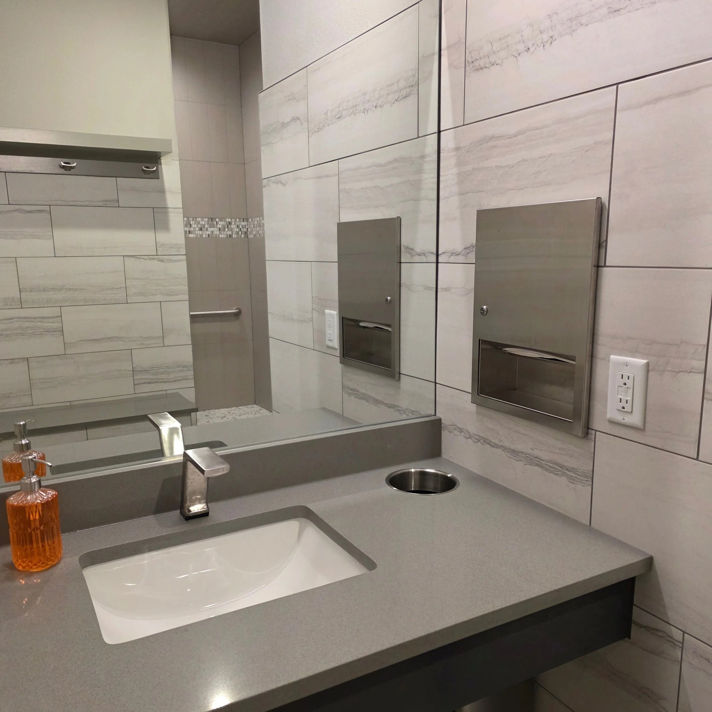 A modern bathroom with a gray countertop, a sink, orange soap dispenser, and a large mirror above. The wall features beige and gray marble tile with horizontal patterns. There are accessed electrical outlets and a stainless steel paper towel or tissue dispenser on the wall.