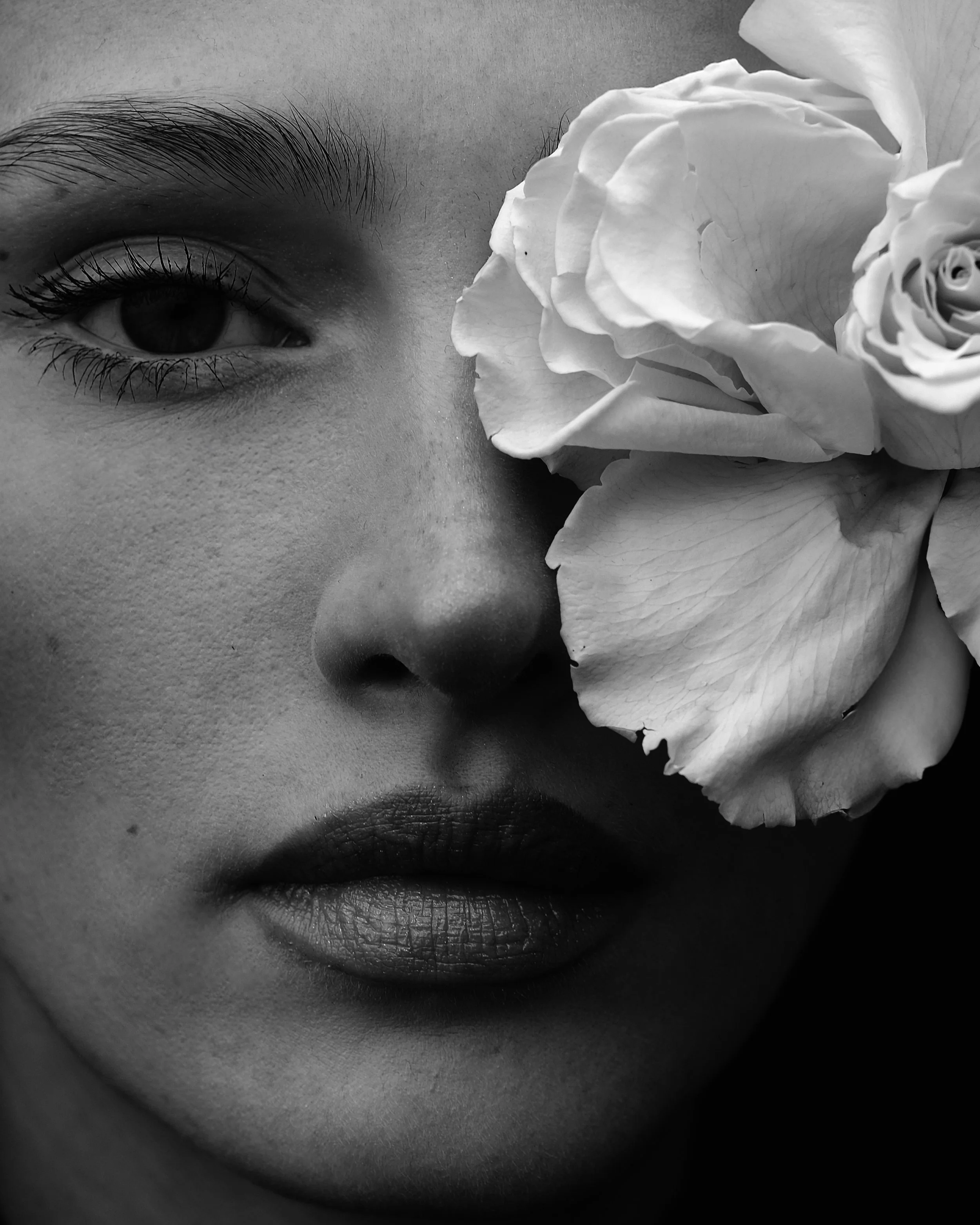 Ritratto in bianco e nero di una donna con un fiore, una rosa, che copre metà volto, mettendo in evidenza l'occhio, il naso e le labbra.