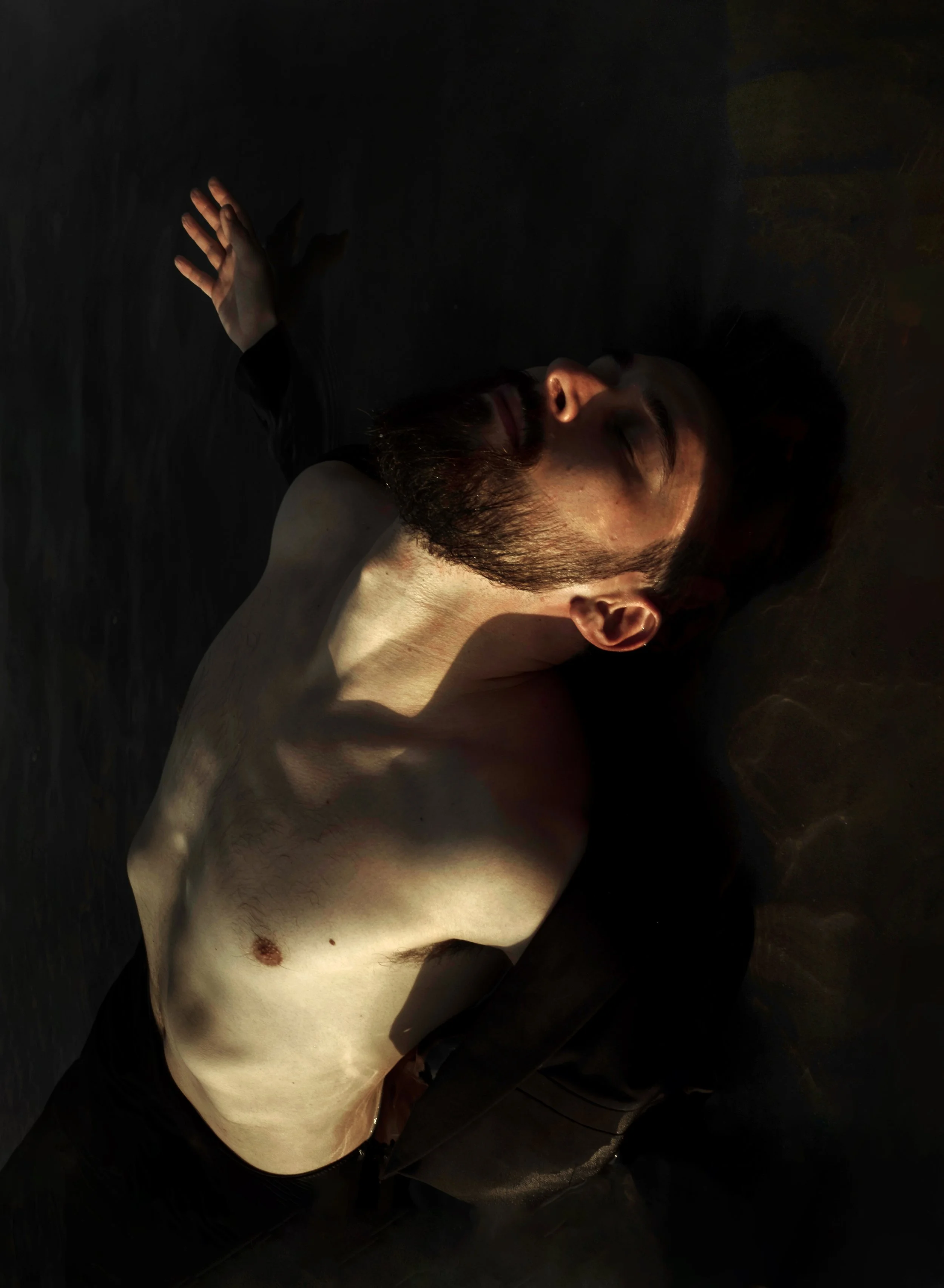 Uomo a torso nudo disteso su una superficie scura, con luce che illumina il volto e il petto.