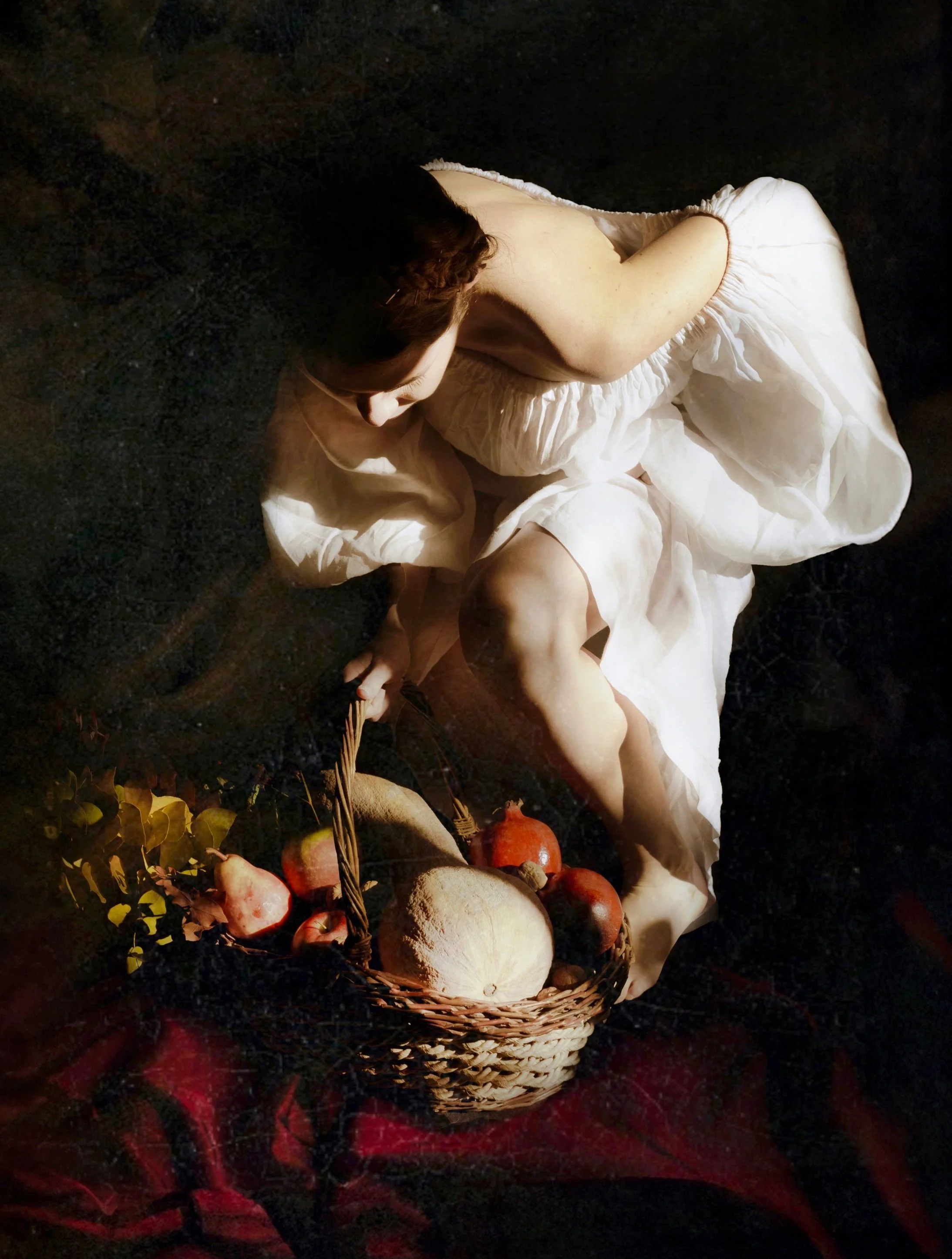Persona distesa su un pavimento scuro, indossa un abito bianco, tiene un cesto con frutta e verdura, tra cui meloni, mele, e pomegranates.