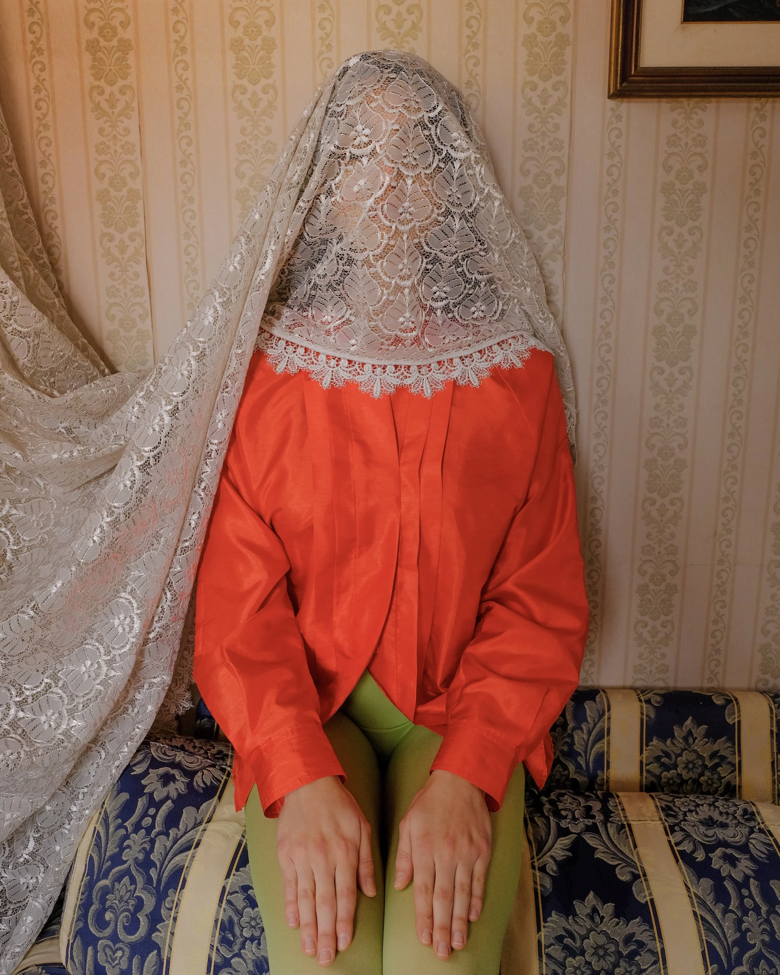 Persona seduta su un divano con un velo di pizzo che copre il viso, indossando una camicia rossa e pantaloni verdi.