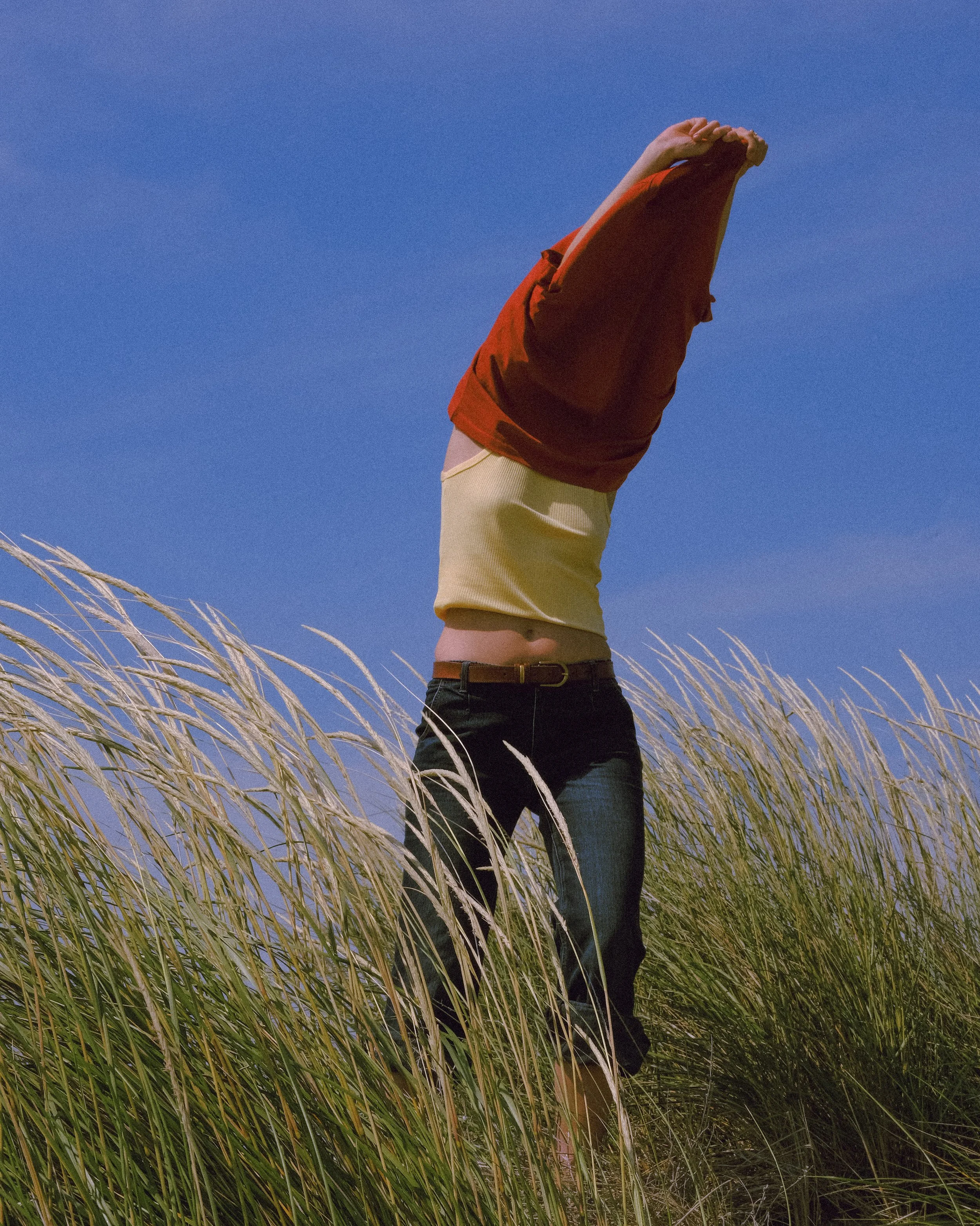 Persona con maglietta beige, pantaloni neri, e giacca rossa che si toglie la giacca in un campo di erba alta sotto un cielo blu.