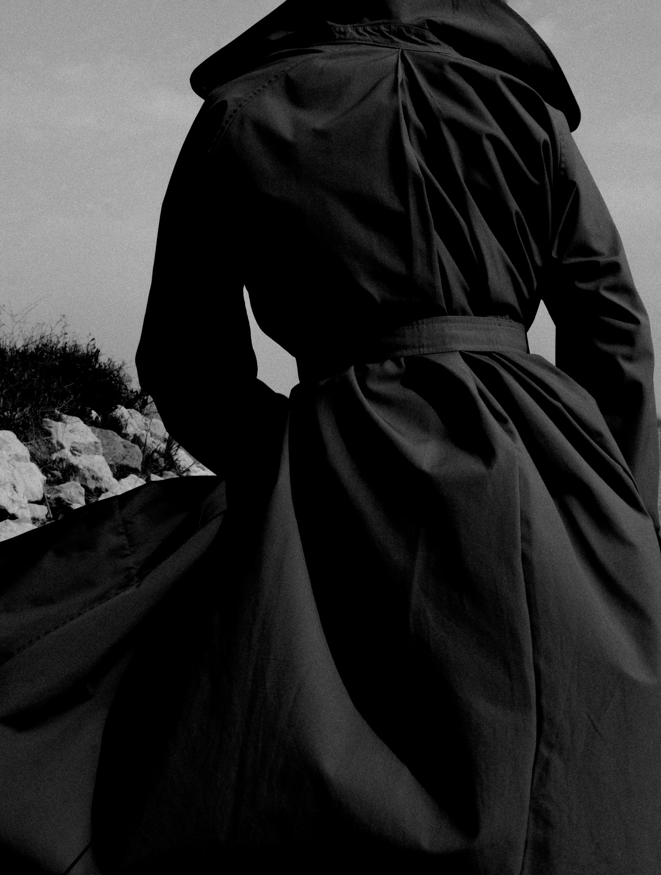 Figura di una persona vestita con un cappotto nero, vista di schiena, che tiene una grande borsa o tessuto in mano, in un ambiente naturale con rocce e vegetazione, in bianco e nero.