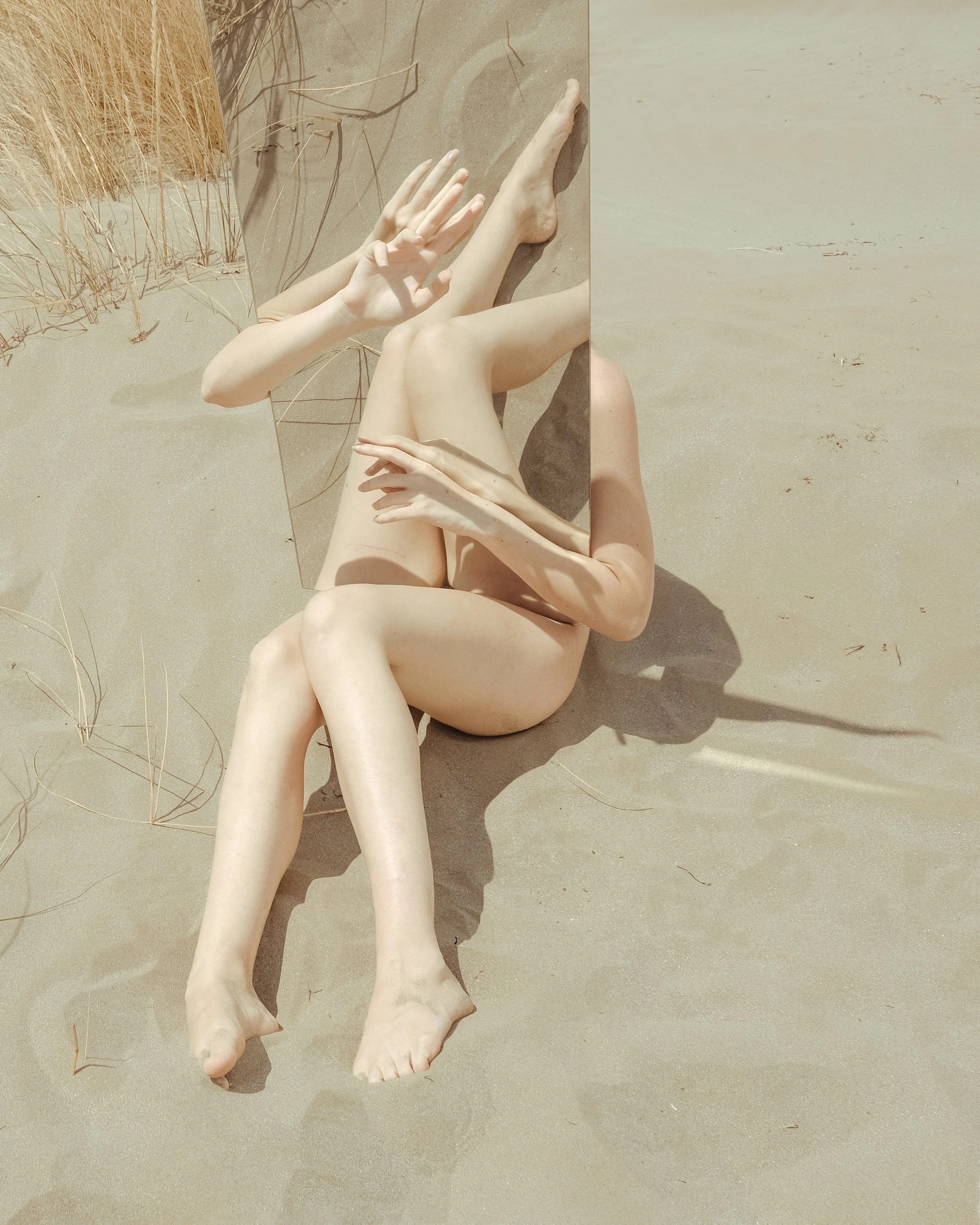 Corpo di una persona disteso sulla sabbia con un grande specchio posizionato in modo che sembri che alcune parti del corpo siano riflesse e ripetute.