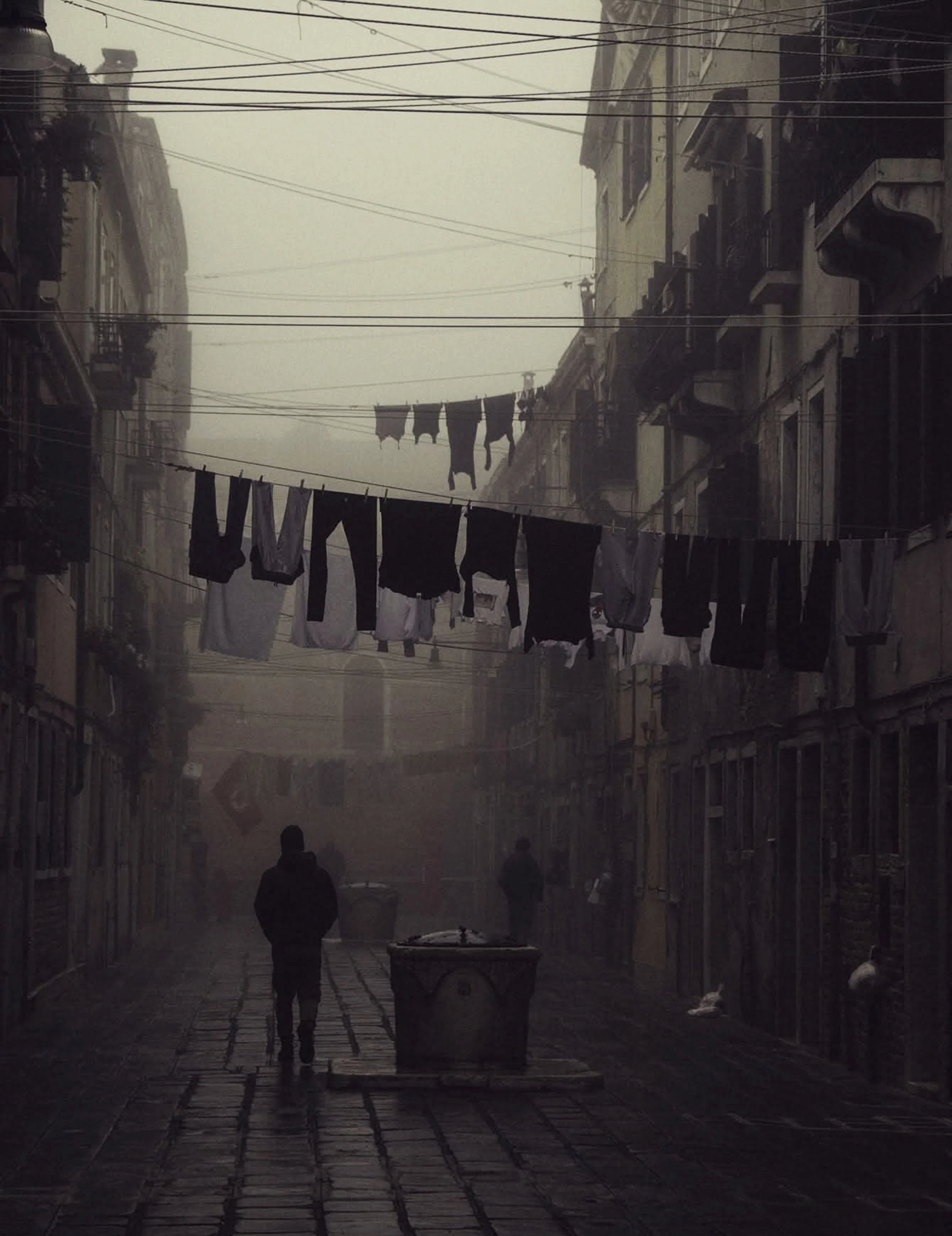 Una strada urbana con edifici vecchi e cavi elettrici sopra, con abiti stesi ad asciugare tra le impalcature. Due persone camminano nella strada, e sembra essere una giornata nebbiosa o piovosa.