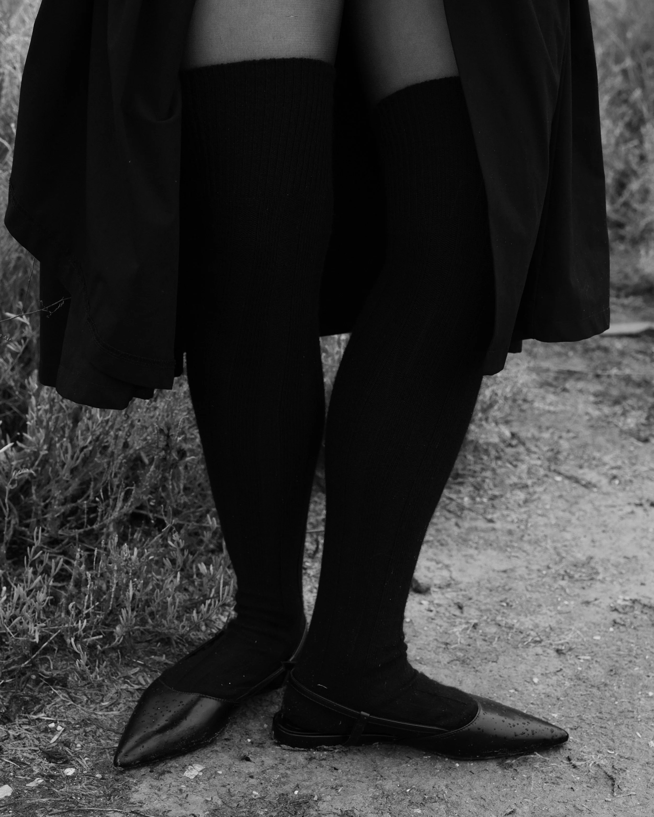 Gambe di una persona con calze lunghe nere e scarpe chiuse su uno sfondo naturale.