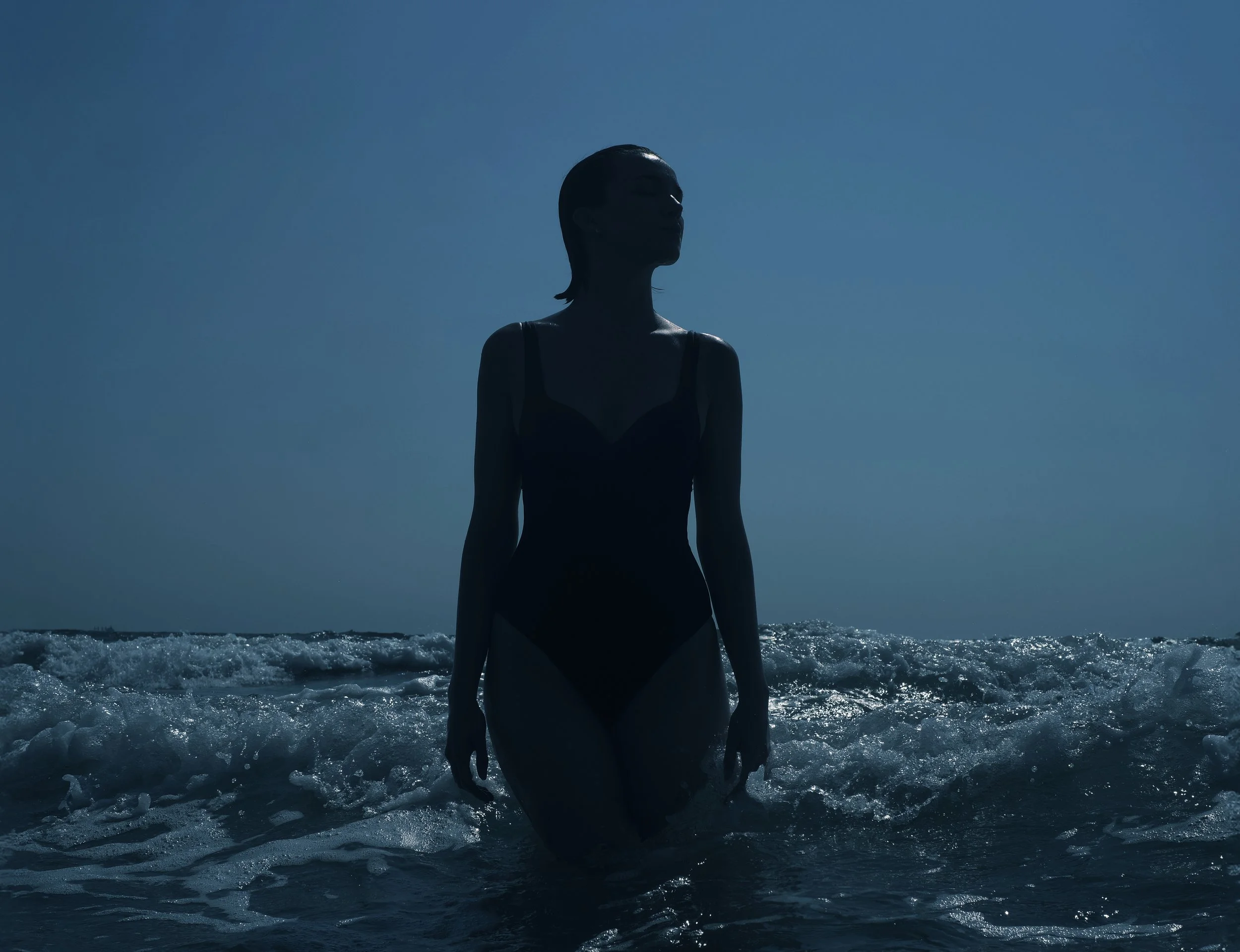 Una donna in costume da bagno si trova in mare con onde che le circondano le gambe, silhouettata contro il cielo chiaro e la luce solare.