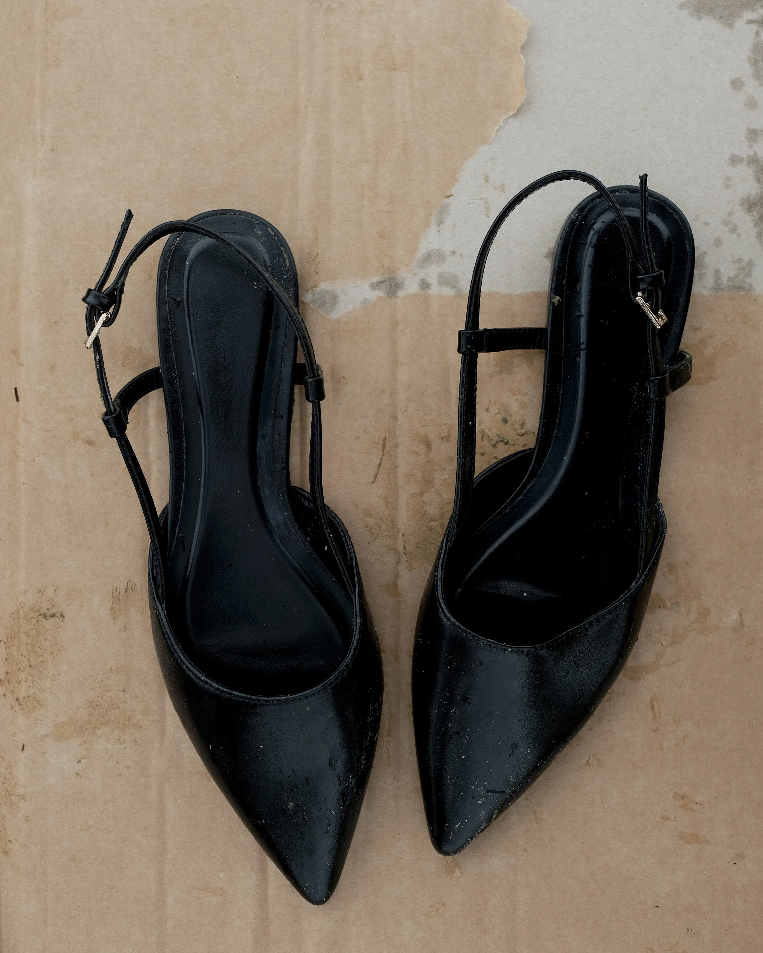 Scarpe nere a punta in pelle con cinturino alla caviglia posizionate su un pavimento in cemento