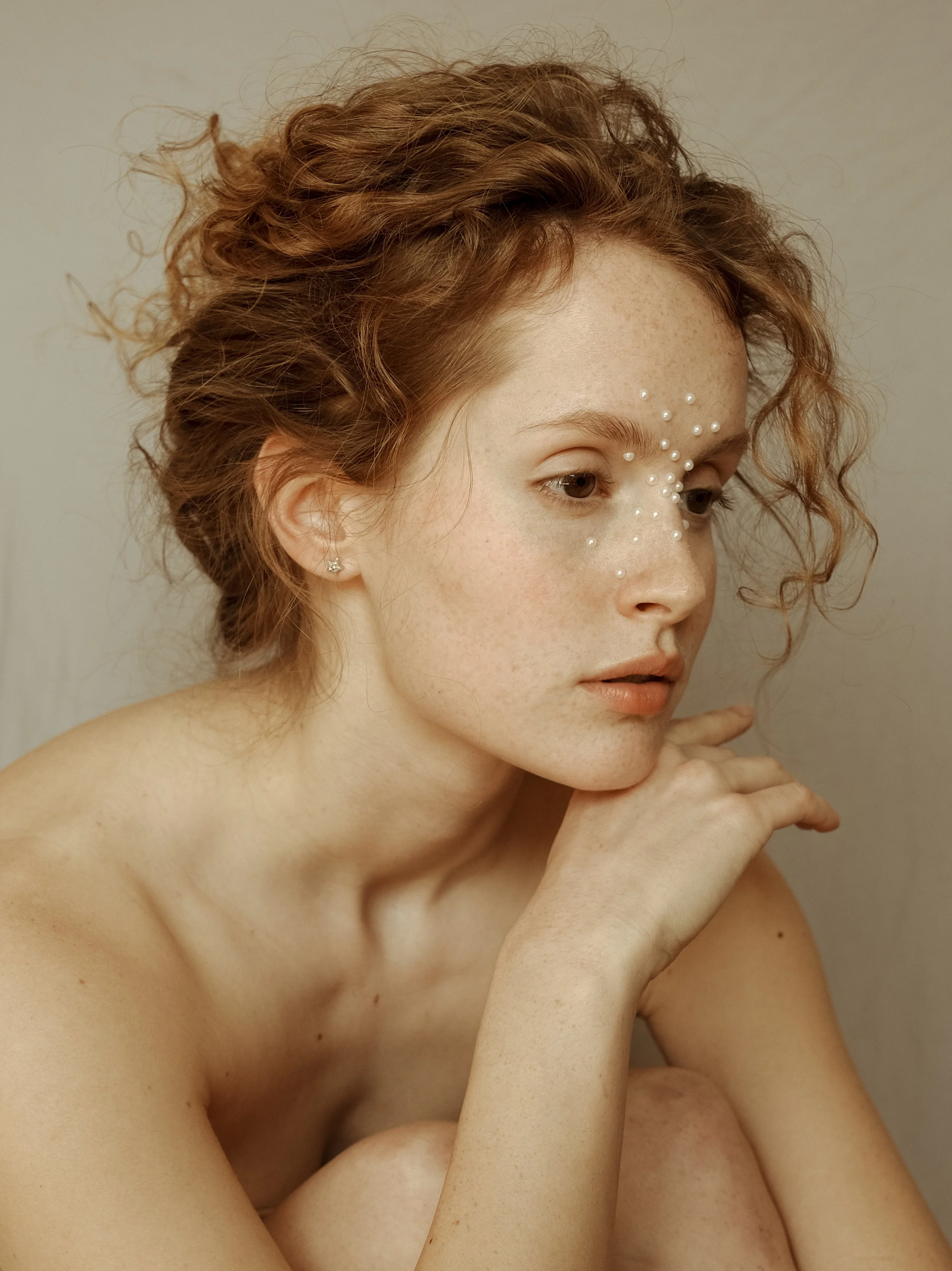 Ritratto di una donna con capelli ricci e perle decorative sul viso in posa pensierosa.