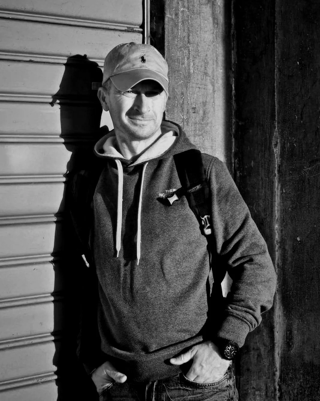 Uomo in abbigliamento casual, con felpa e cappellino, in piedi vicino a una serranda, fotografato in bianco e nero.