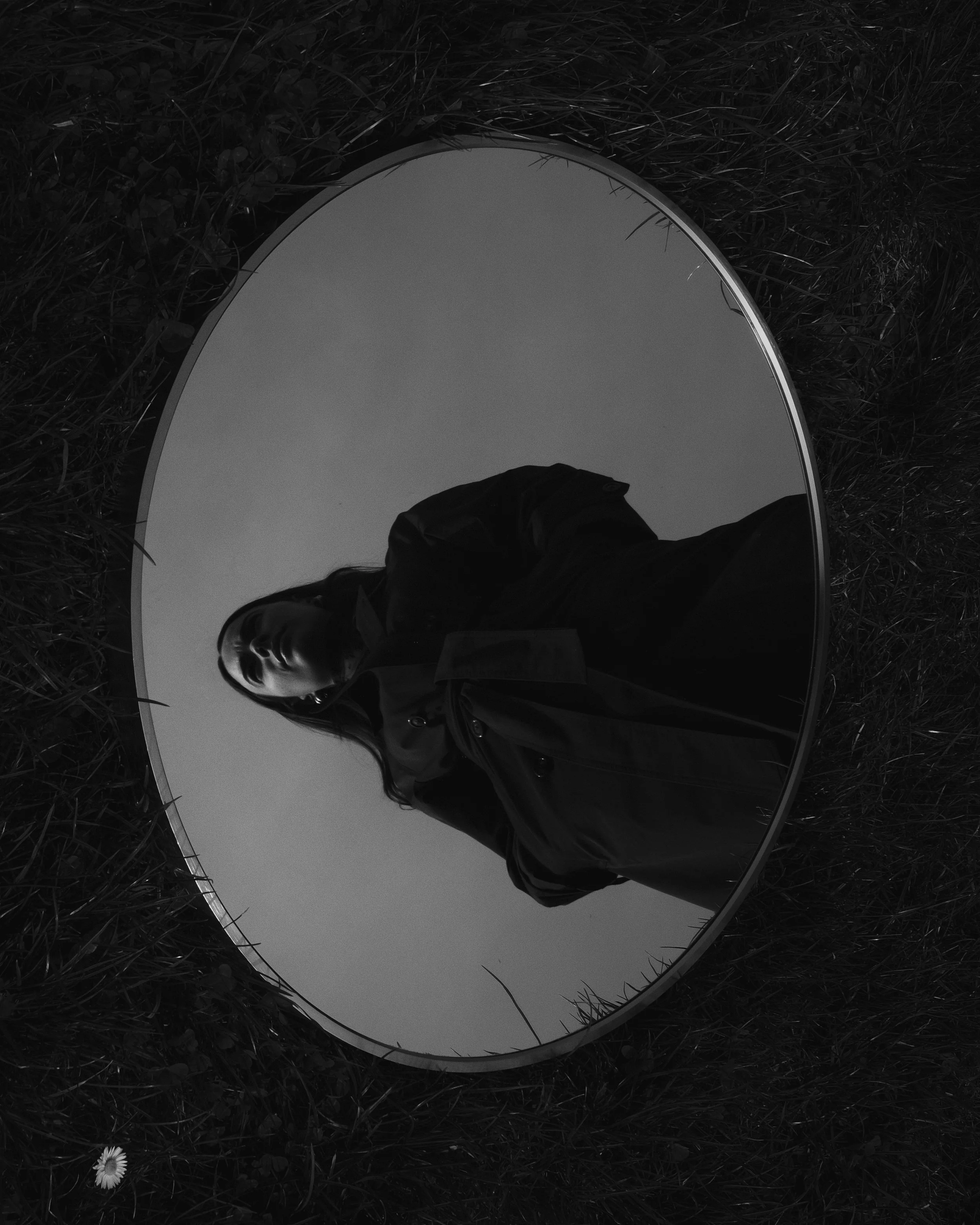 Riflesso di una donna in uno specchio rotondo posto su terra con erba, in un ambiente esterno.