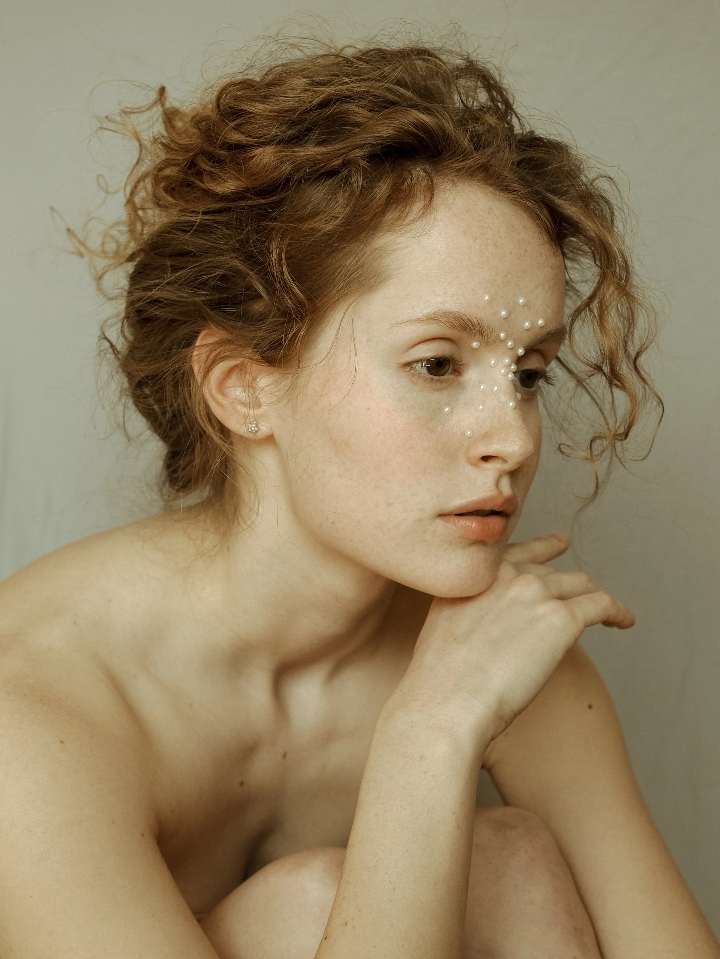 Ritratto di una donna con capelli ricci e rossi, con decorazioni di perline sulla faccia, che posa con il mento appoggiato sulla mano.