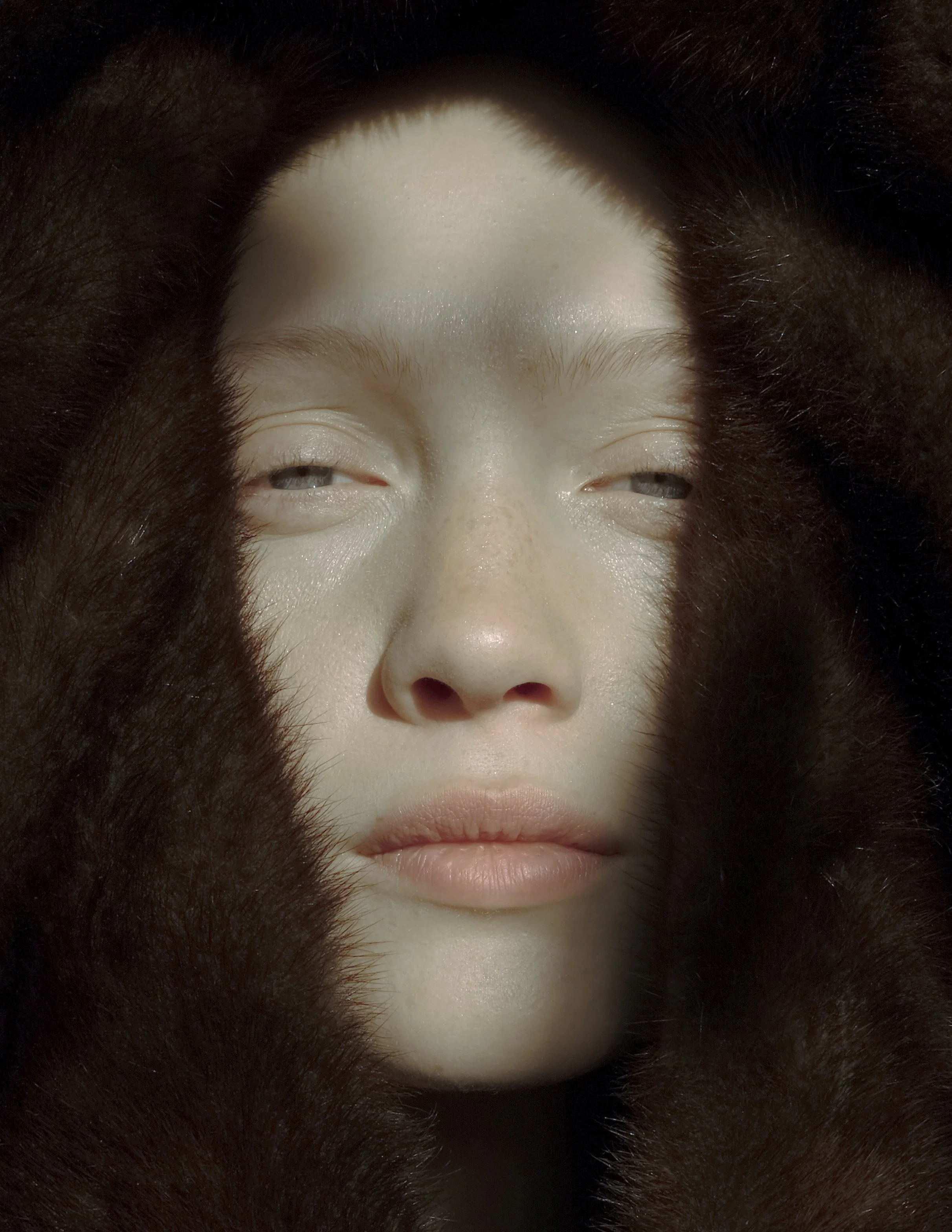 Primo piano di un volto incorniciato da un tessuto peloso scuro, con occhi semiaperti e luce che crea ombre.