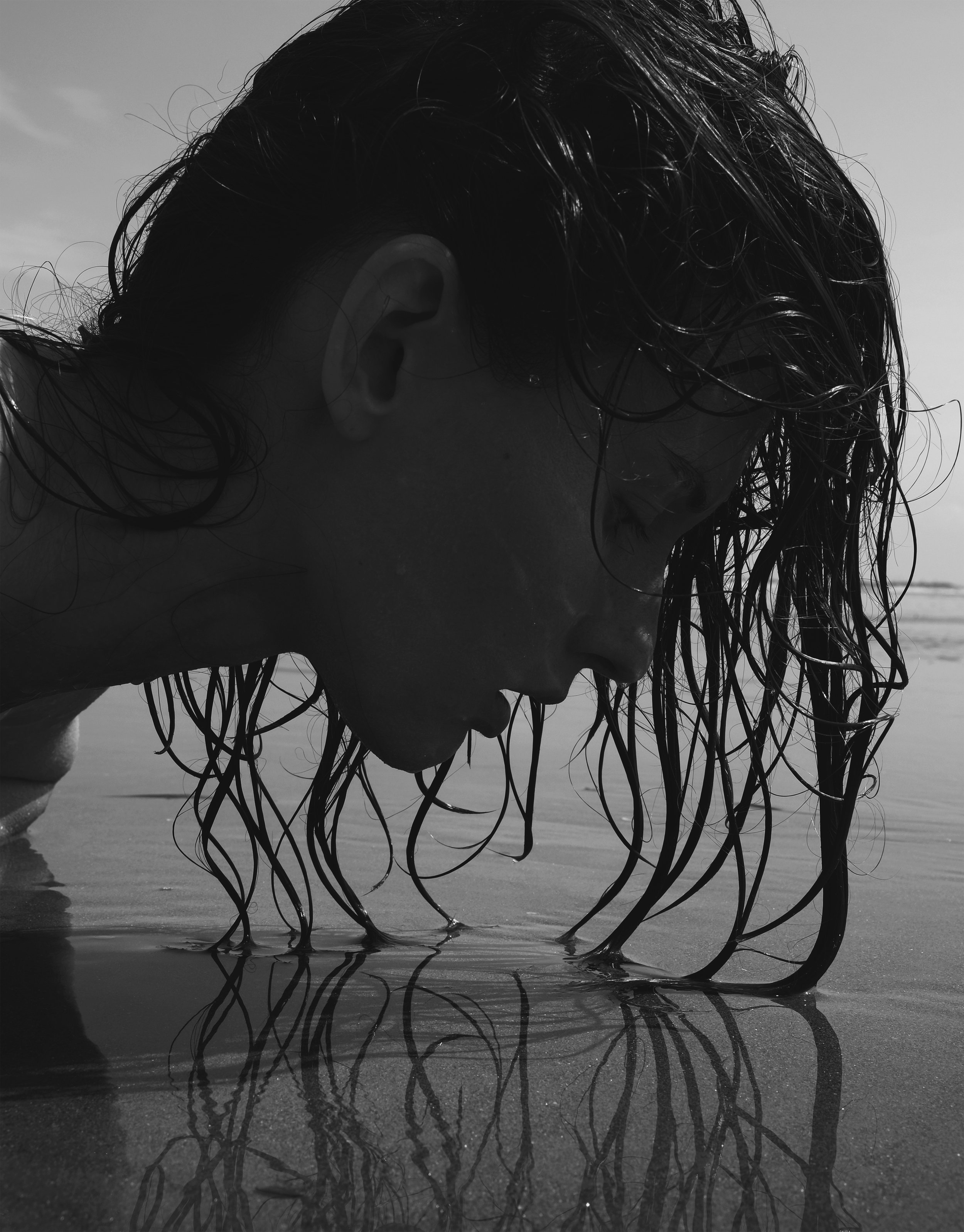 Ragazza con capelli bagnati che si china sull'acqua sulla spiaggia in bianco e nero.