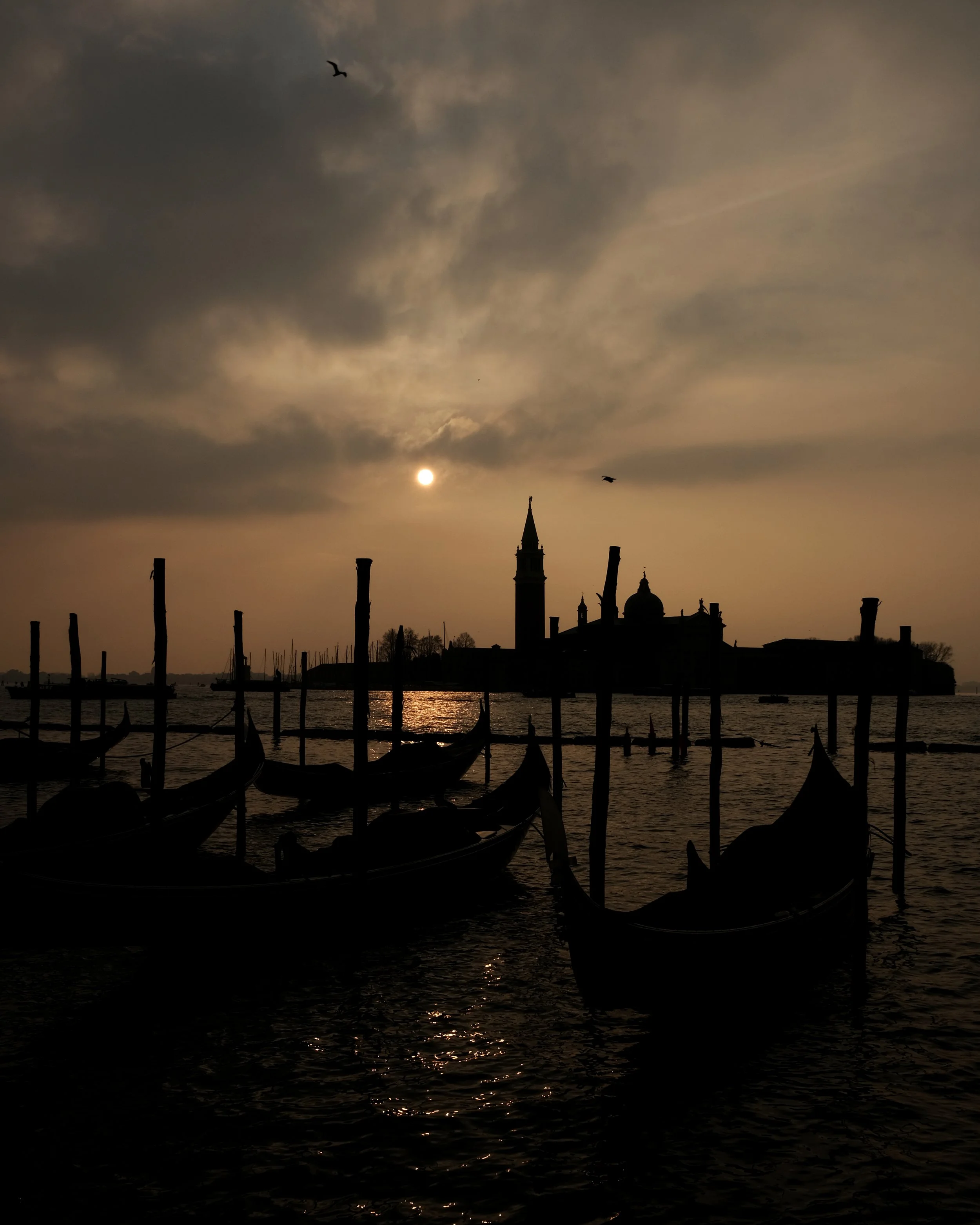 Veduta notturna di Venezia con gondole in primo piano e il Campanile di San Marco sullo sfondo, sotto un cielo nuvoloso con il sole che tramonta.