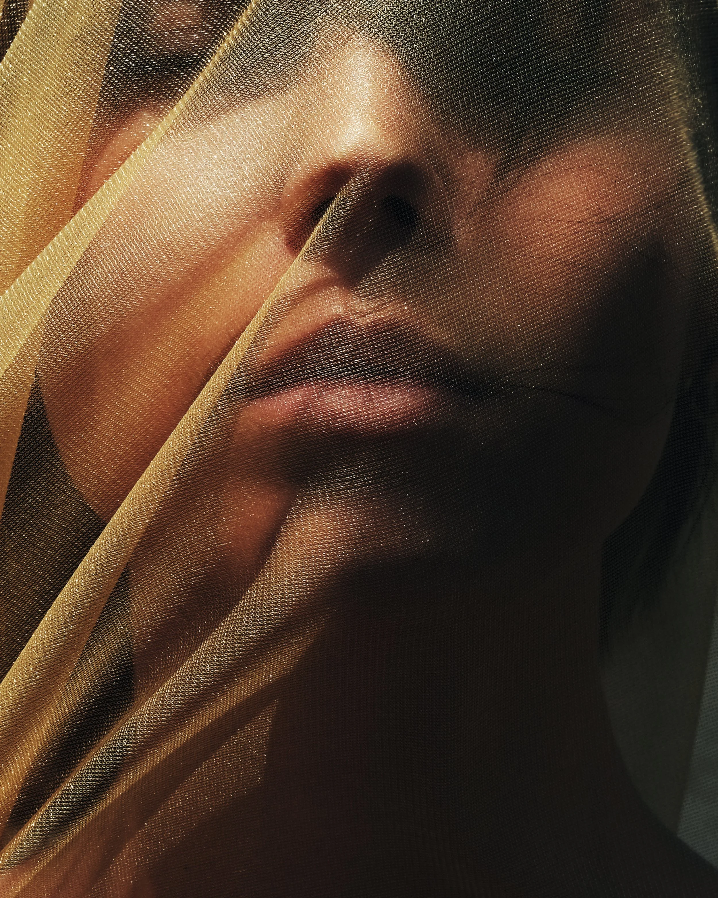 Ritratto di una donna con il volto parzialmente coperto da un velo di tessuto dorato, che lascia intravedere le labbra, il naso e una parte del viso.