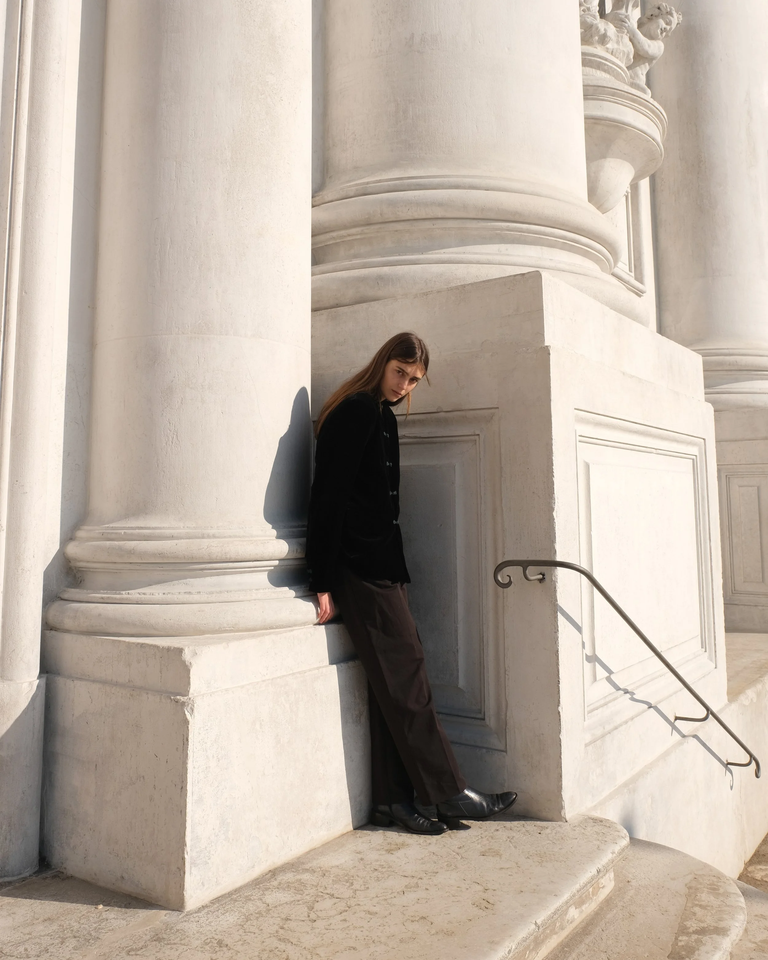 Persona in piedi accanto a una grande colonna bianca con scale a lato, indossando abbigliamento scuro.
