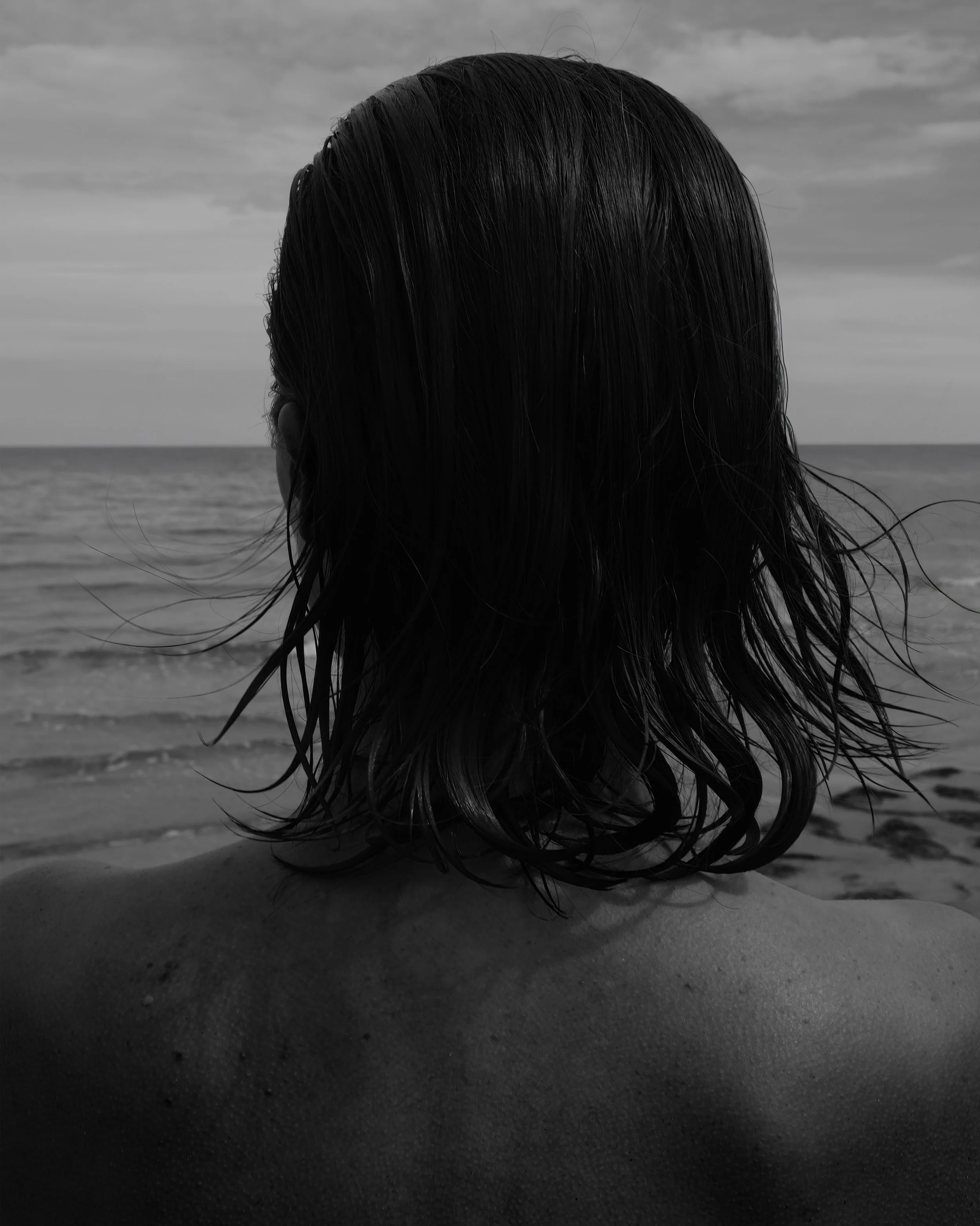 Vista di una persona con i capelli bagnati, vista dalla schiena, sulla spiaggia con il mare sullo sfondo.