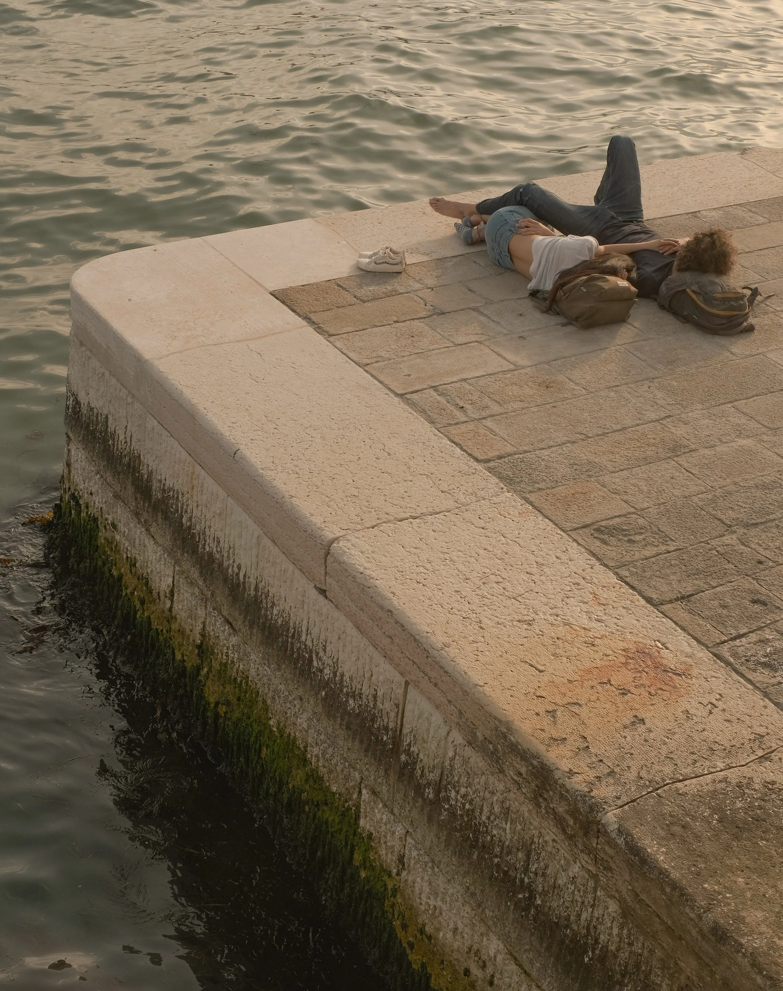 Due persone sdraiate su una banchina vicino all'acqua, con uno zaino e una scarpa bianca a fianco. La superficie della banchina è in pietra.