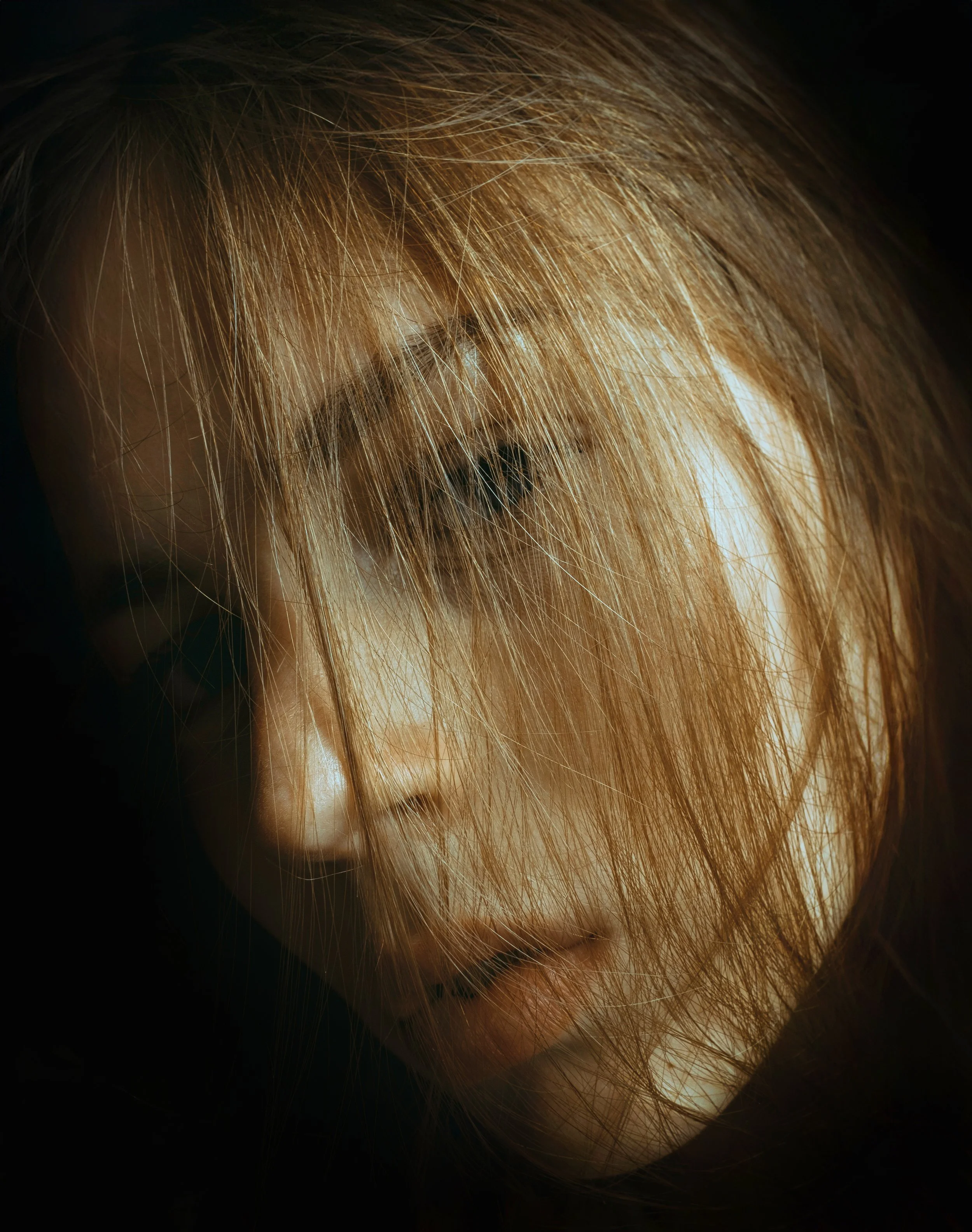 Ritratto ravvicinato di una persona con capelli lunghi castani, viso parzialmente coperto dai capelli, illuminazione fioca.