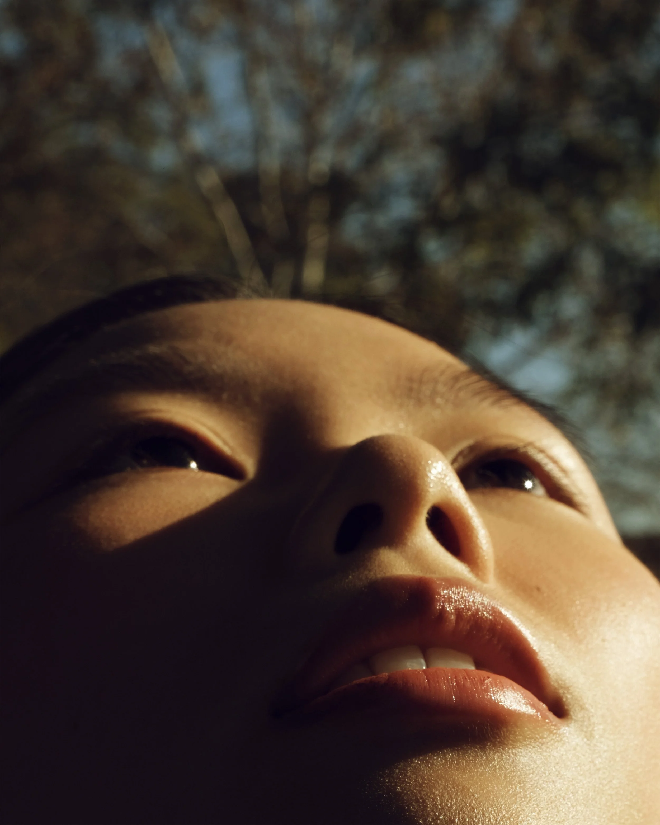 Primo piano di un volto di una persona, vista dal basso, con uno sfondo di cielo e alberi.