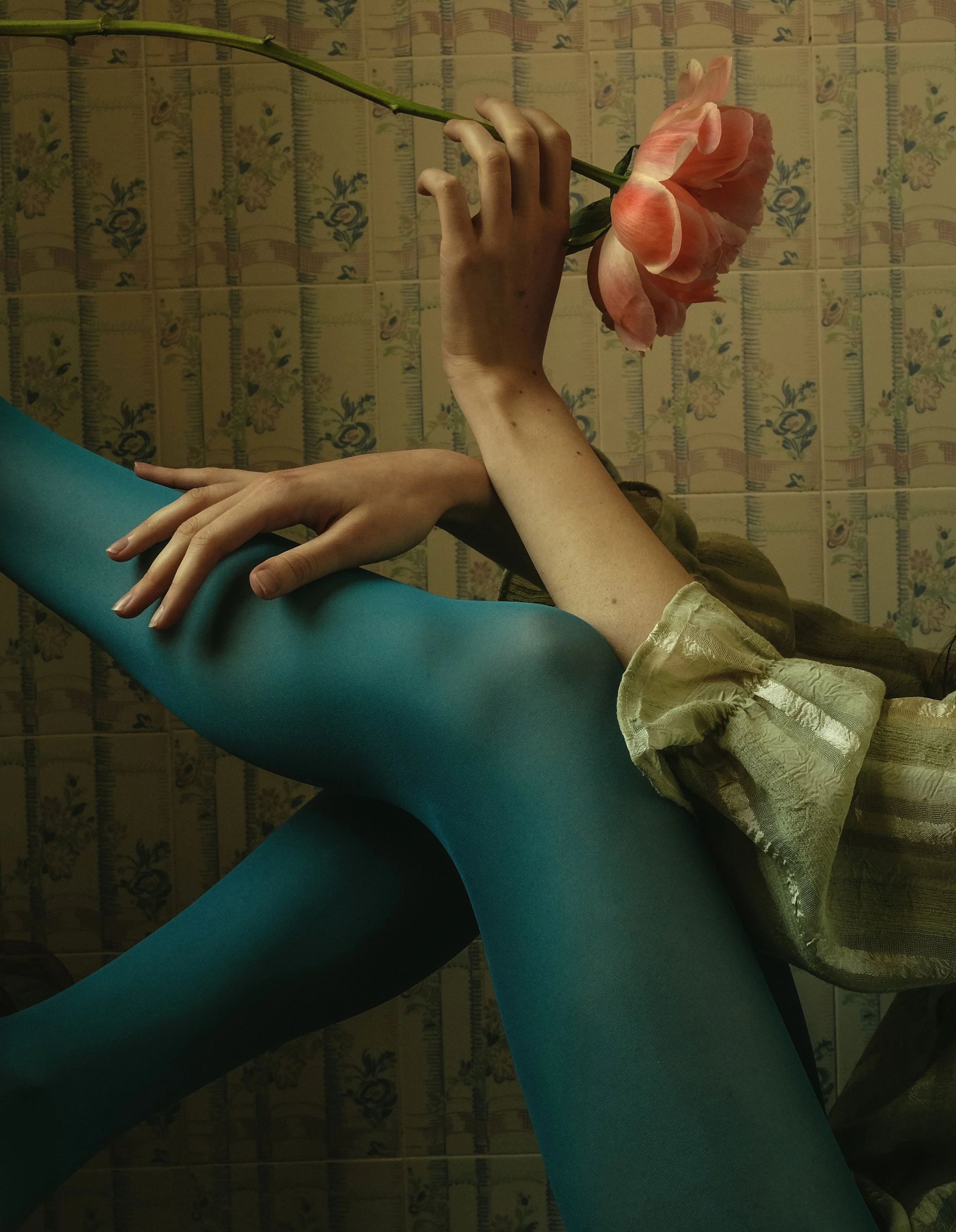 Persona con collant verdi e camicetta, tenendo un grande fiore rosa, sfondo floreale.