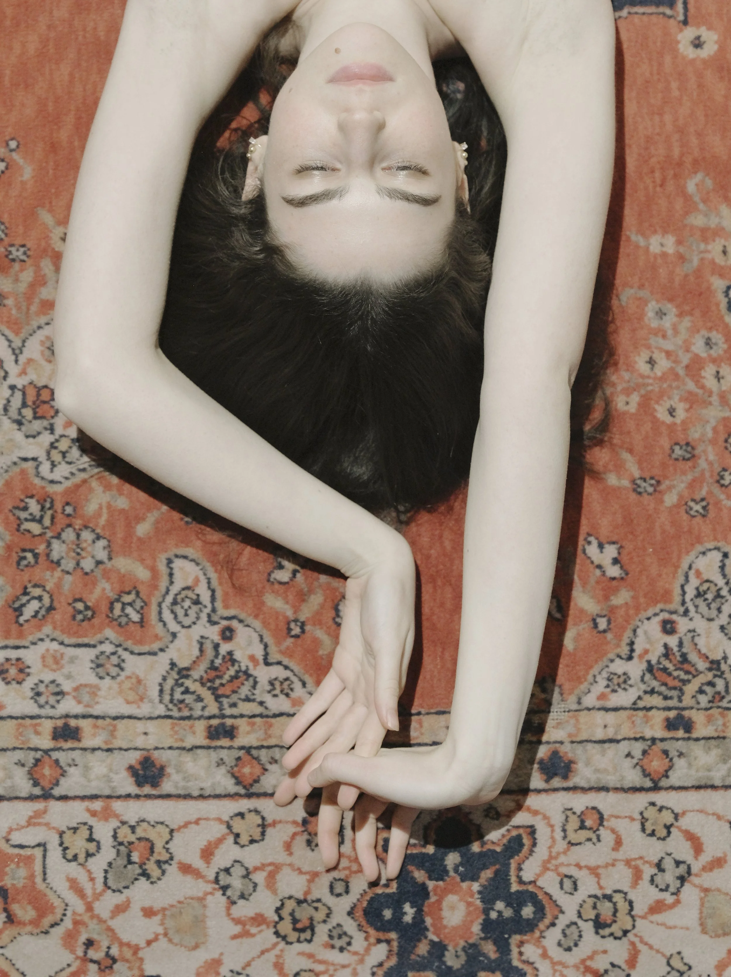 Ragazza con pelle molto chiara e capelli scuri, con le mani incrociate sopra la testa, sdraiata su un tappeto arancione.