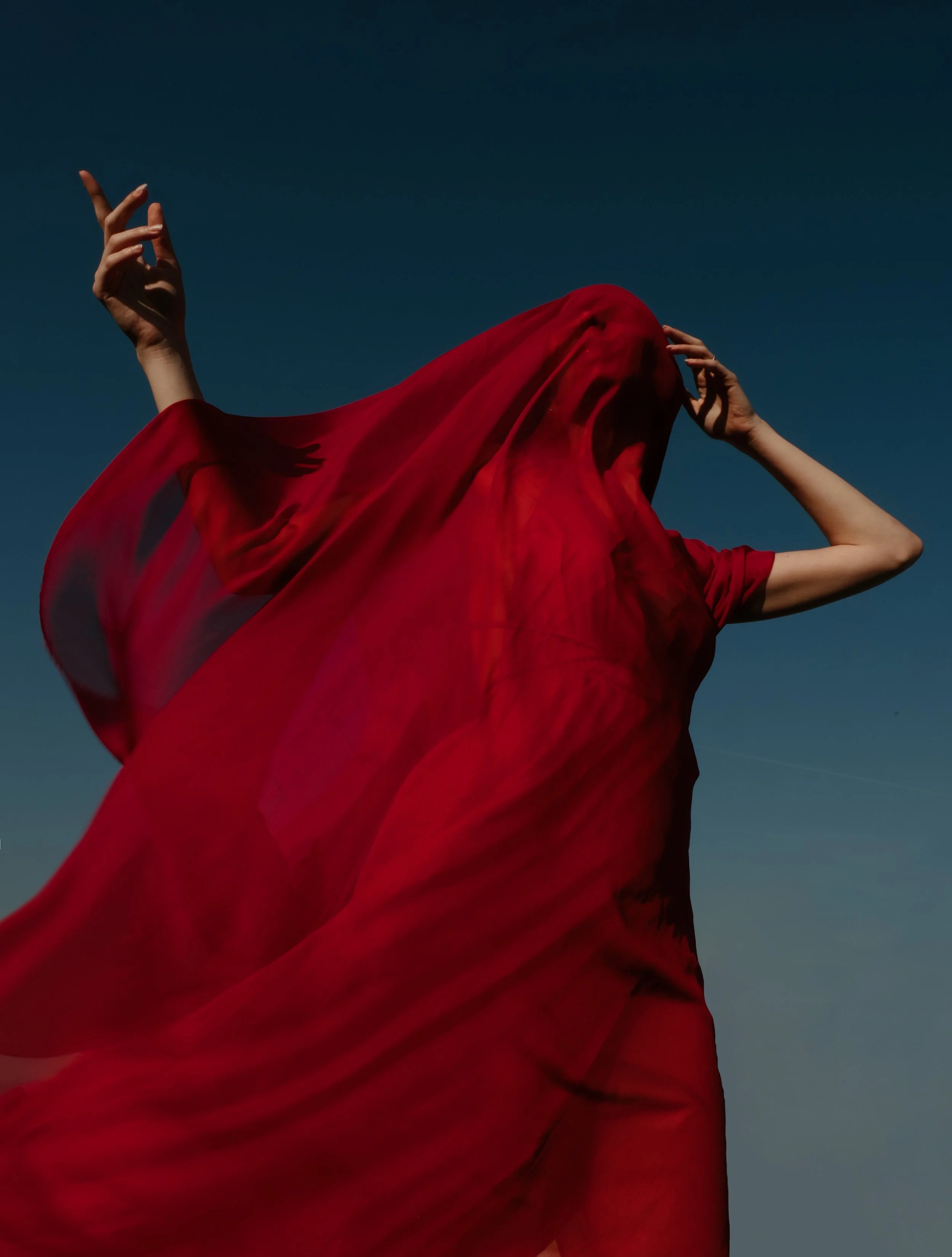 Persona avvolta in un tessuto rosso trasparente, con il cielo blu sullo sfondo.