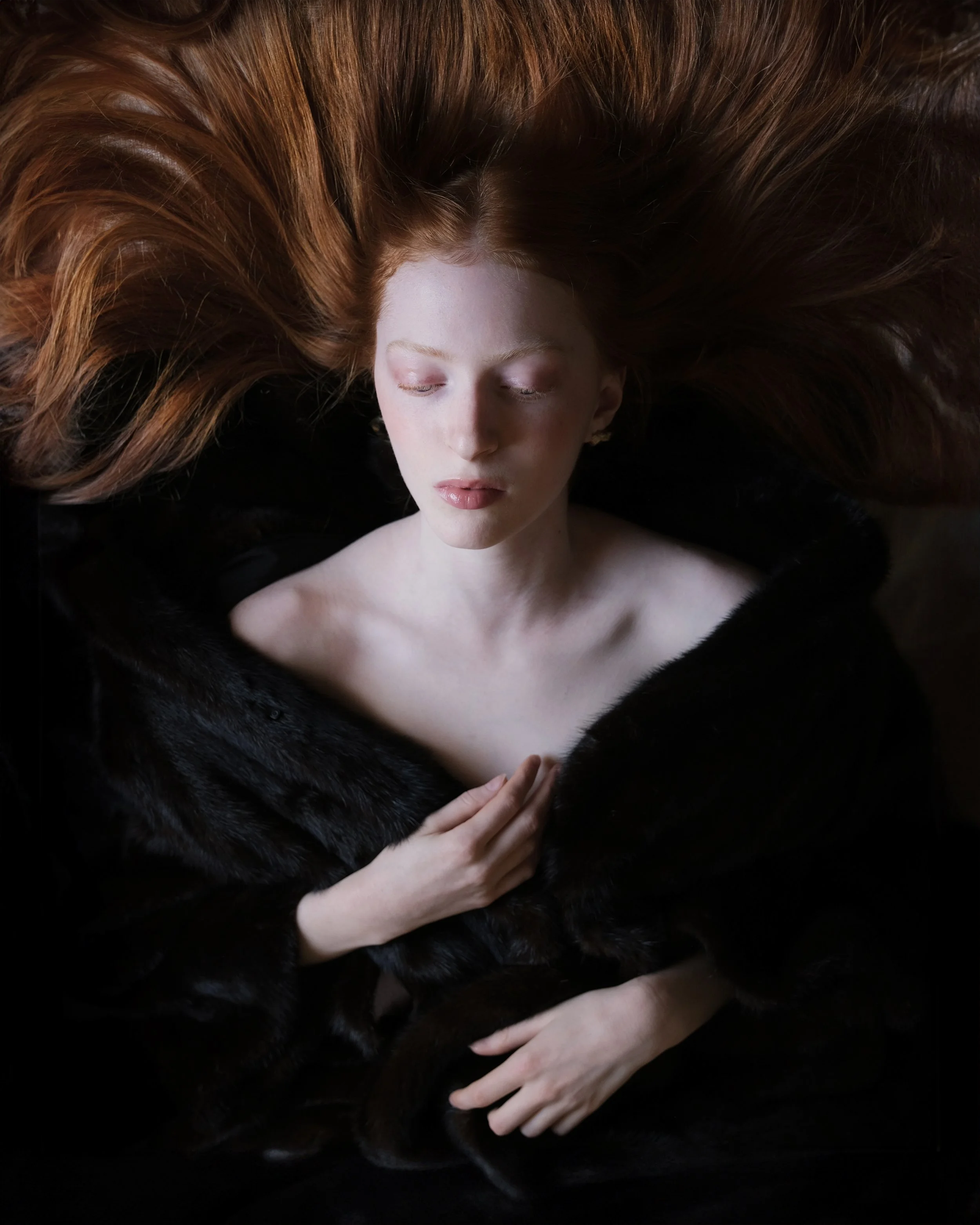 Una donna con i lunghi capelli rossi chiusi gli occhi, indossa un cappotto di pelliccia nera e si trova in un ambiente scuro.