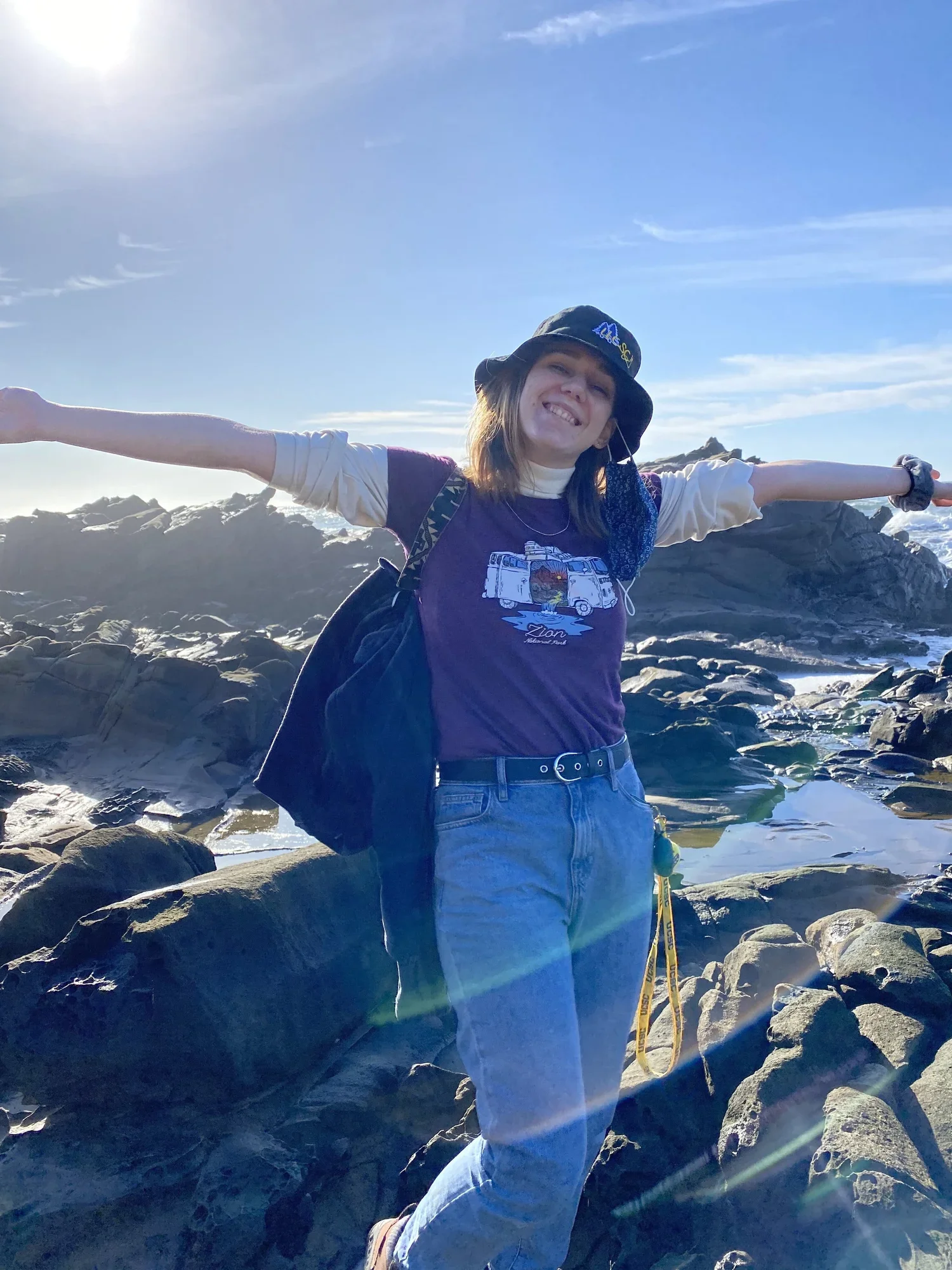 A woman standing on rocks near the water with arms outstretched, wearing a purple graphic T-shirt, blue jeans, a bucket hat, and a backpack, smiling with the ocean, rocks, and sky in the background.