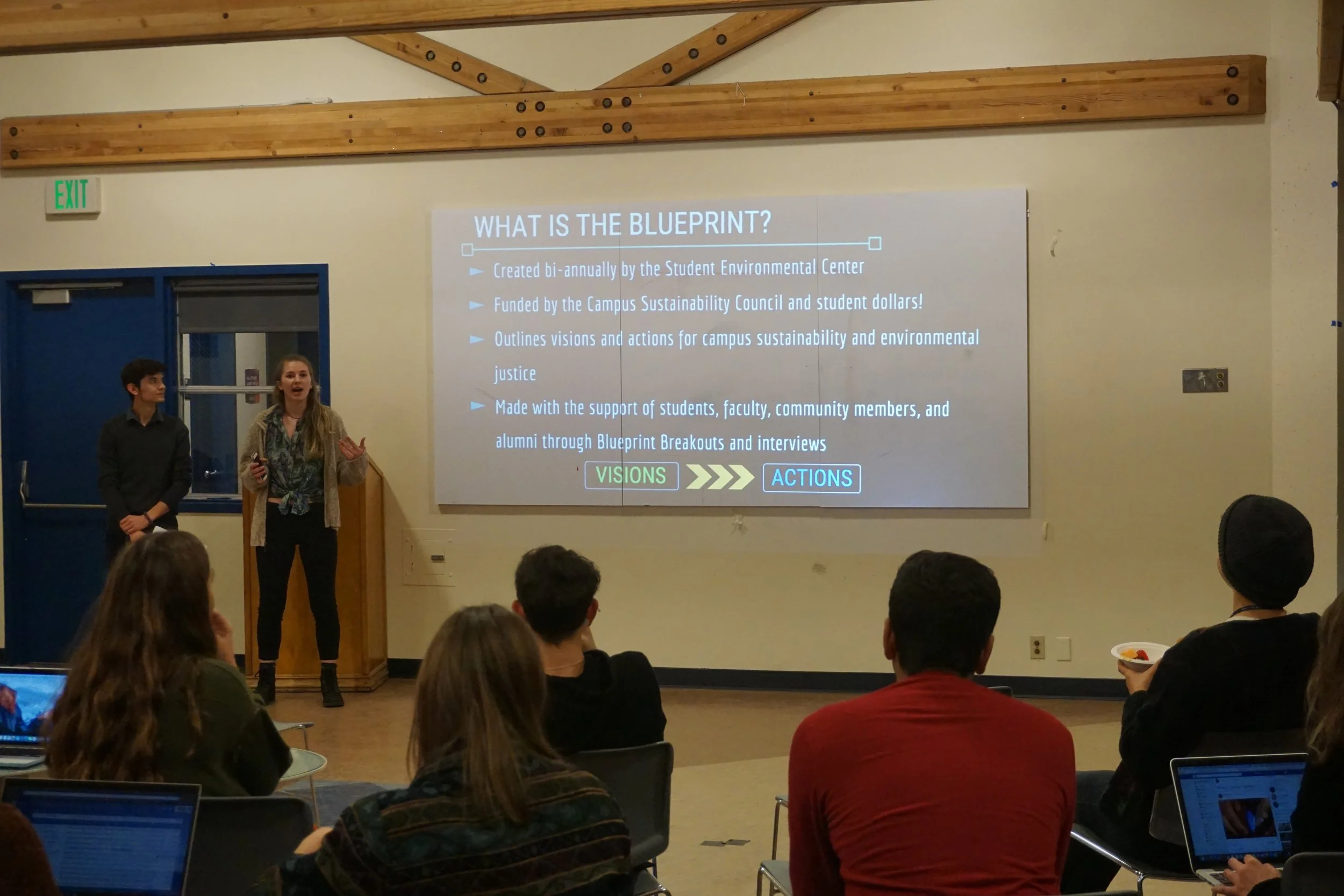 Two young presenters, a male and a female, standing at the front of a room with a projection screen displaying information about a blueprint. The audience is seated, some taking notes on laptops.