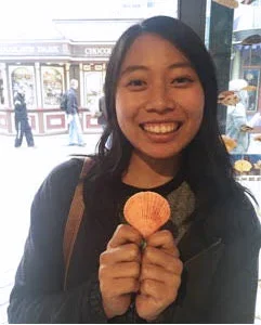 Smiling woman holding a cookie in a cafe with large windows and city street outside.