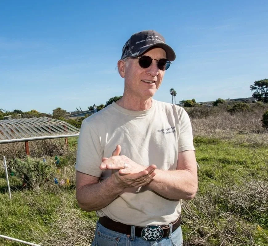 An elderly man with sunglasses, a baseball cap, and a beige t-shirt stands outdoors in a grassy area, smiling and gesturing with his hands.