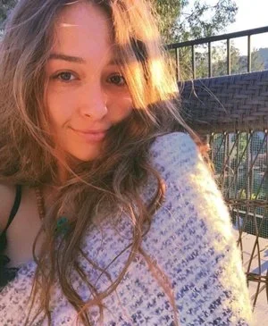 A young woman with long wavy hair taking a selfie outside on a sunny day, with a railing and trees in the background.