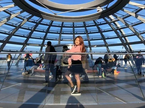 Young woman in pink sweater and black skirt smiling inside a glass dome structure with people around and blue sky above.