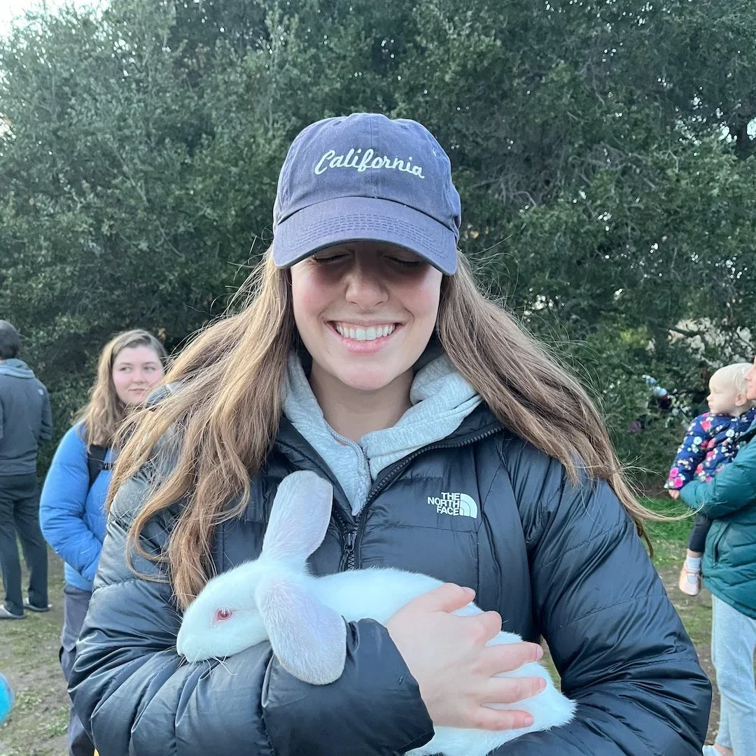 Young woman smiling and holding a white rabbit outdoors, wearing a gray cap with 'California' embroidered and a black North Face jacket, surrounded by other people and trees.