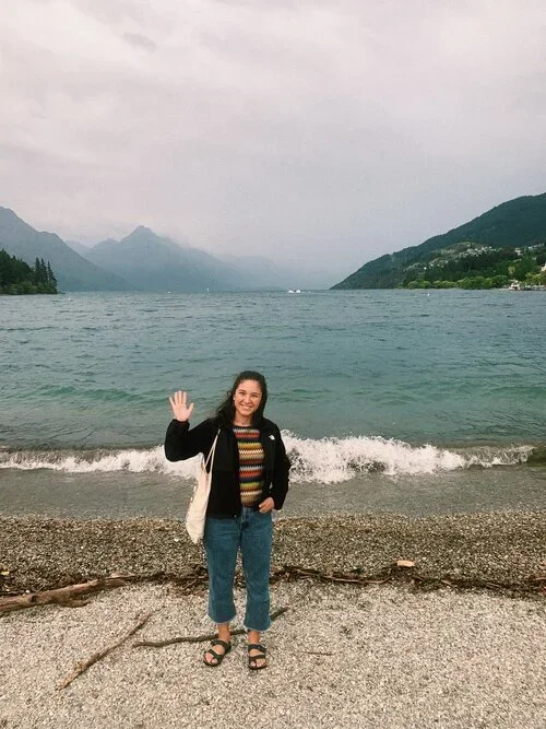 A woman standing on a beach, waving, with a large lake, mountains, and cloudy sky in the background.
