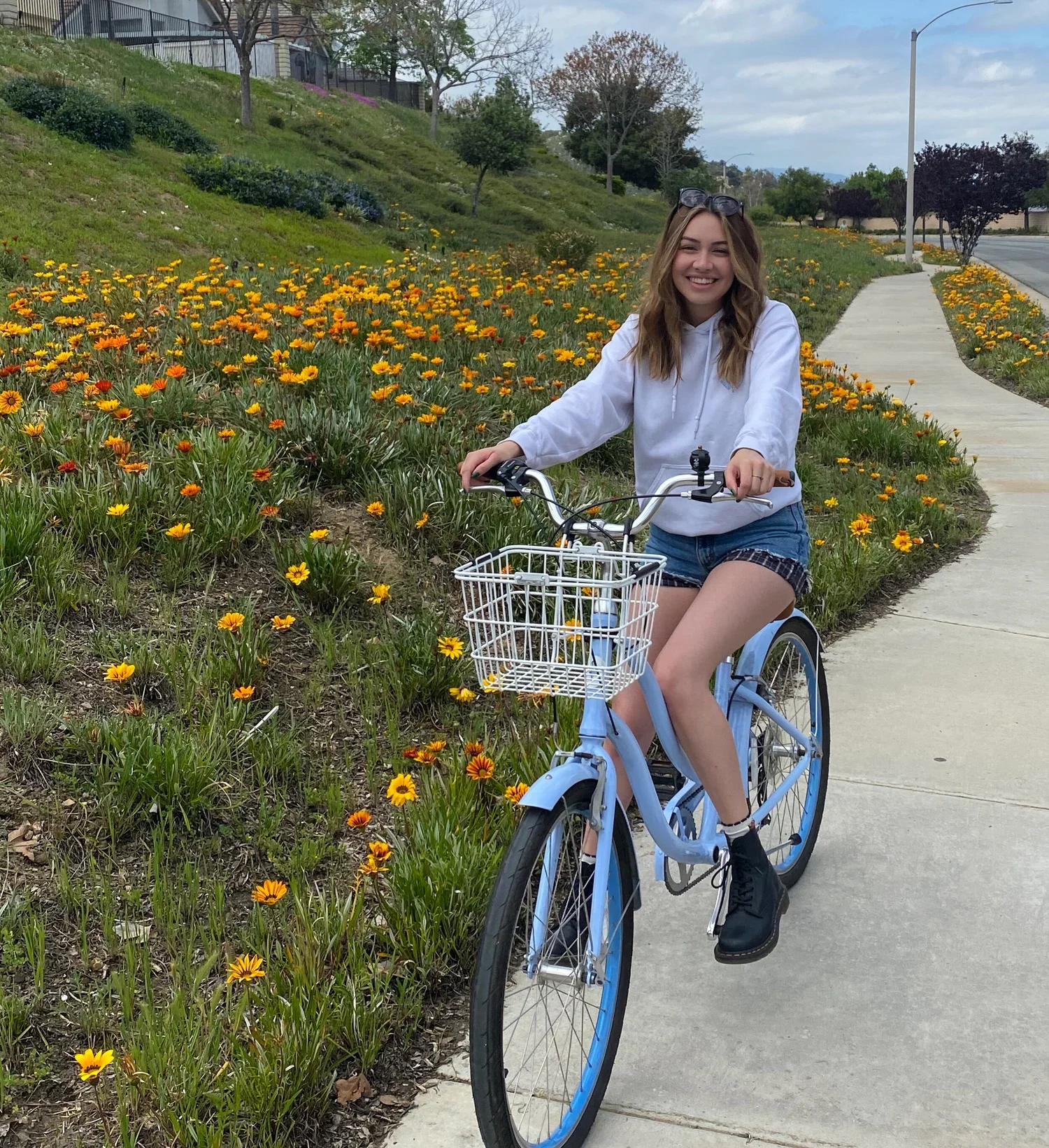 A young woman with long wavy hair riding a light blue bicycle along a sidewalk beside a flower bed with orange and yellow flowers. She is smiling, wearing a white hoodie, shorts, black boots, and sunglasses on her head.