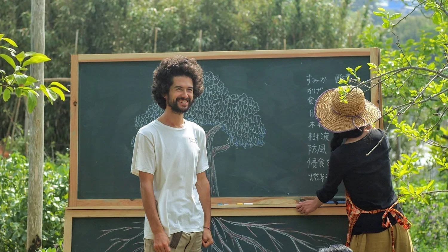 A man with curly hair and a beard smiling outdoors next to a woman writing on a blackboard that has a tree drawing on it. The woman is wearing a large straw hat and a black shirt with an apron. The scene is surrounded by greenery.