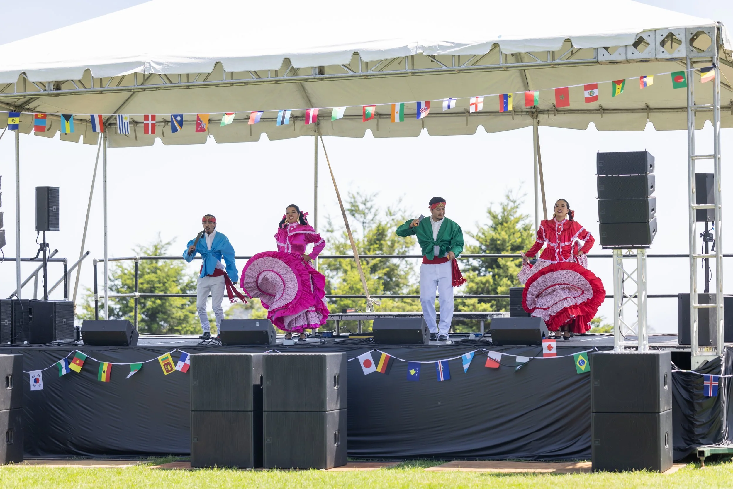 Performers dancing on an outdoor stage decorated with international flags, dressed in colorful traditional Mexican dresses and shirts.