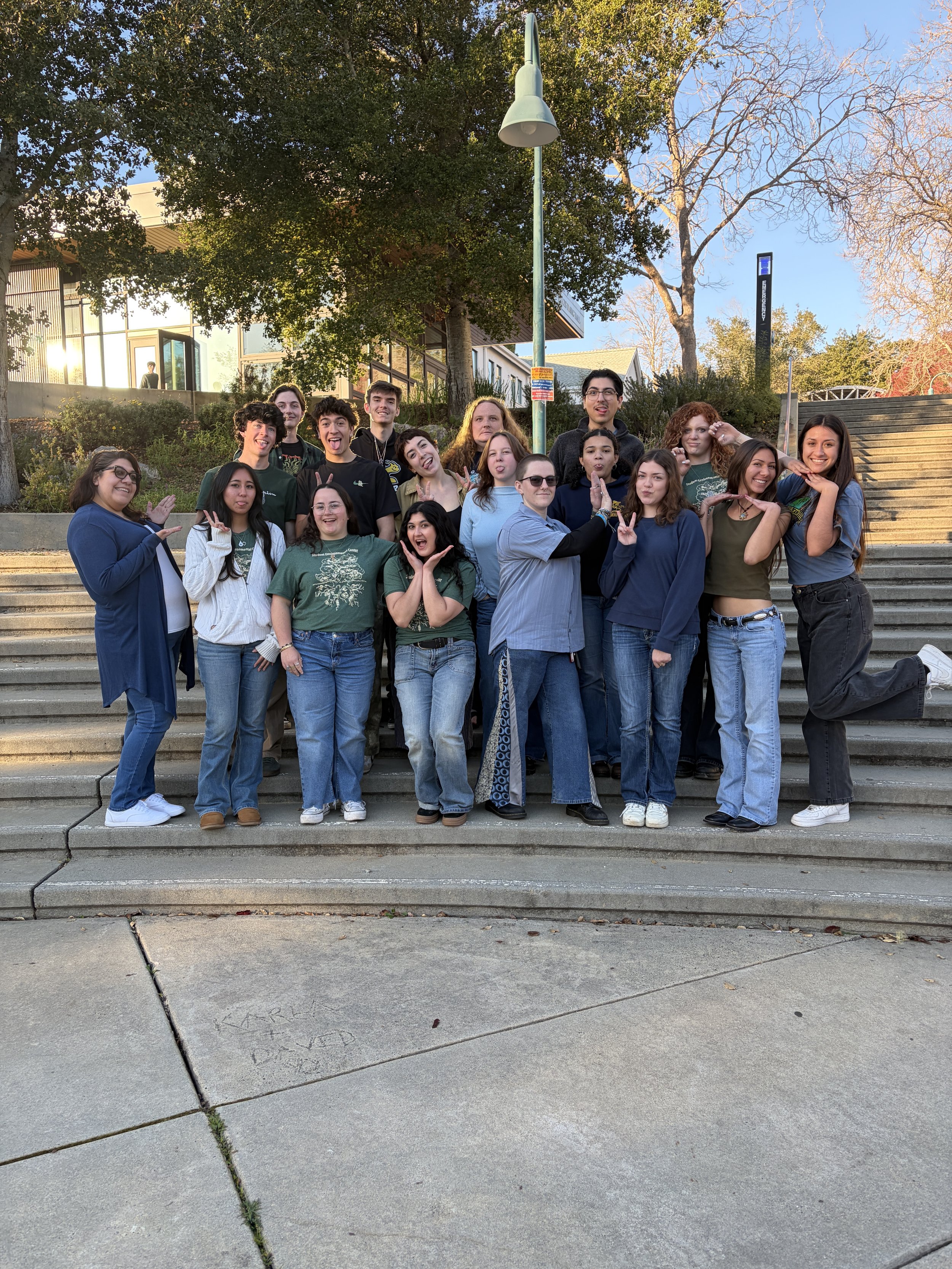 Group of young people standing on outdoor steps, posing for a photo, with some making playful gestures, in a park-like setting with trees and buildings in the background during daylight.