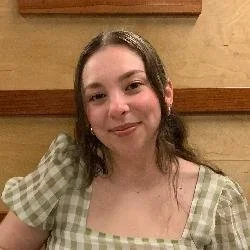 A young woman with shoulder-length brown hair smiling at the camera, wearing a green patterned top, sitting indoors with a wooden panel background.