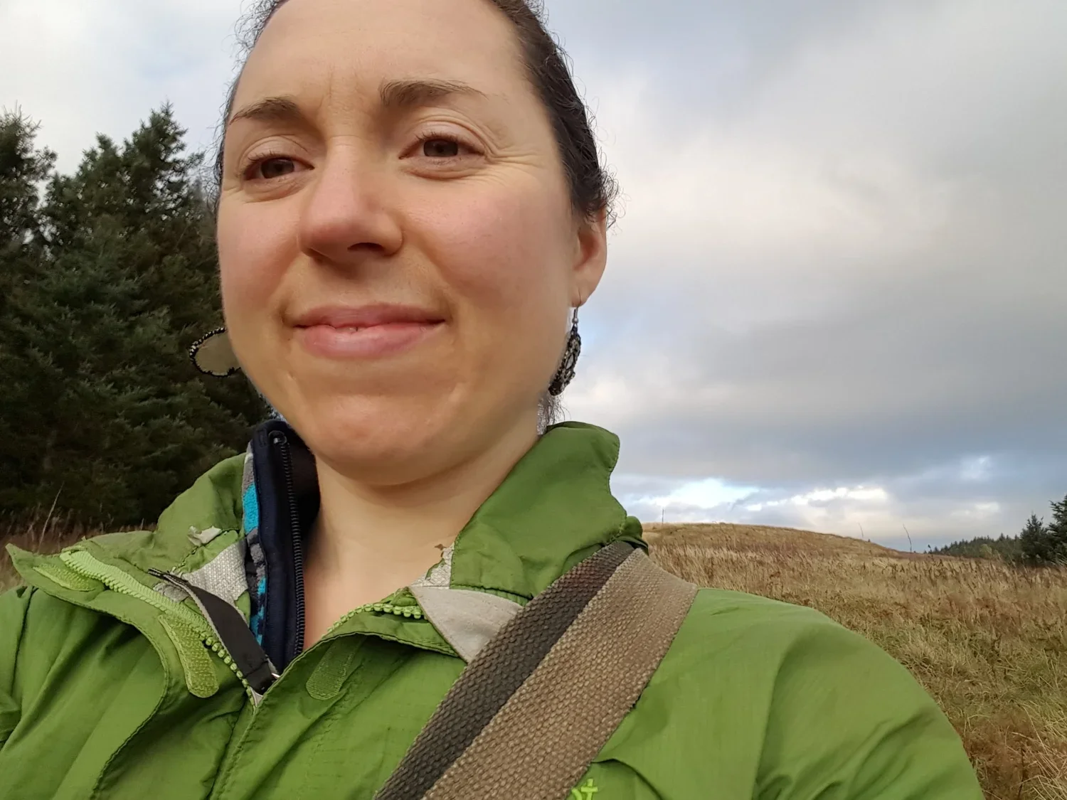 Close-up of a woman taking a selfie outdoors with trees and hills in the background, wearing a green jacket and earrings.