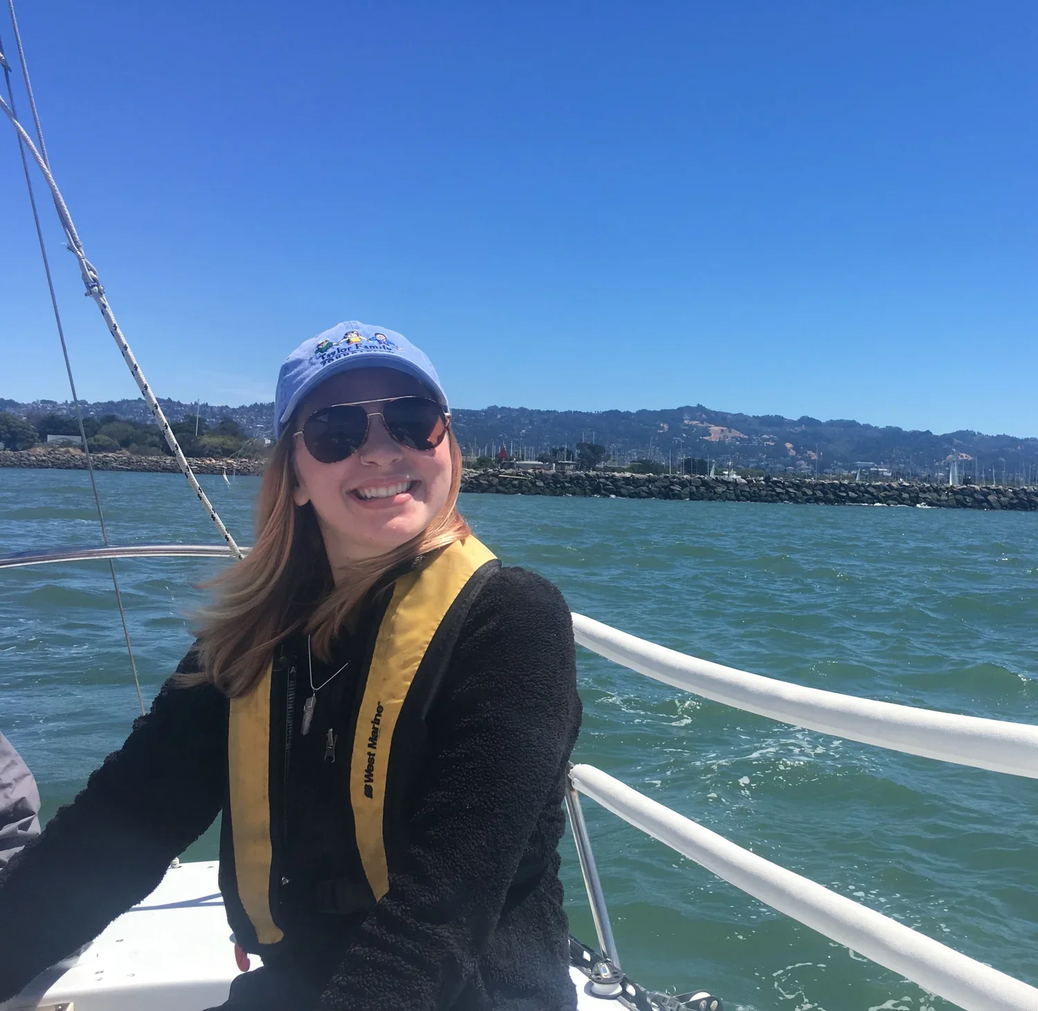 Woman with sunglasses and a baseball cap smiling while sailing on a boat in a bay with hills and sailboats in the background.