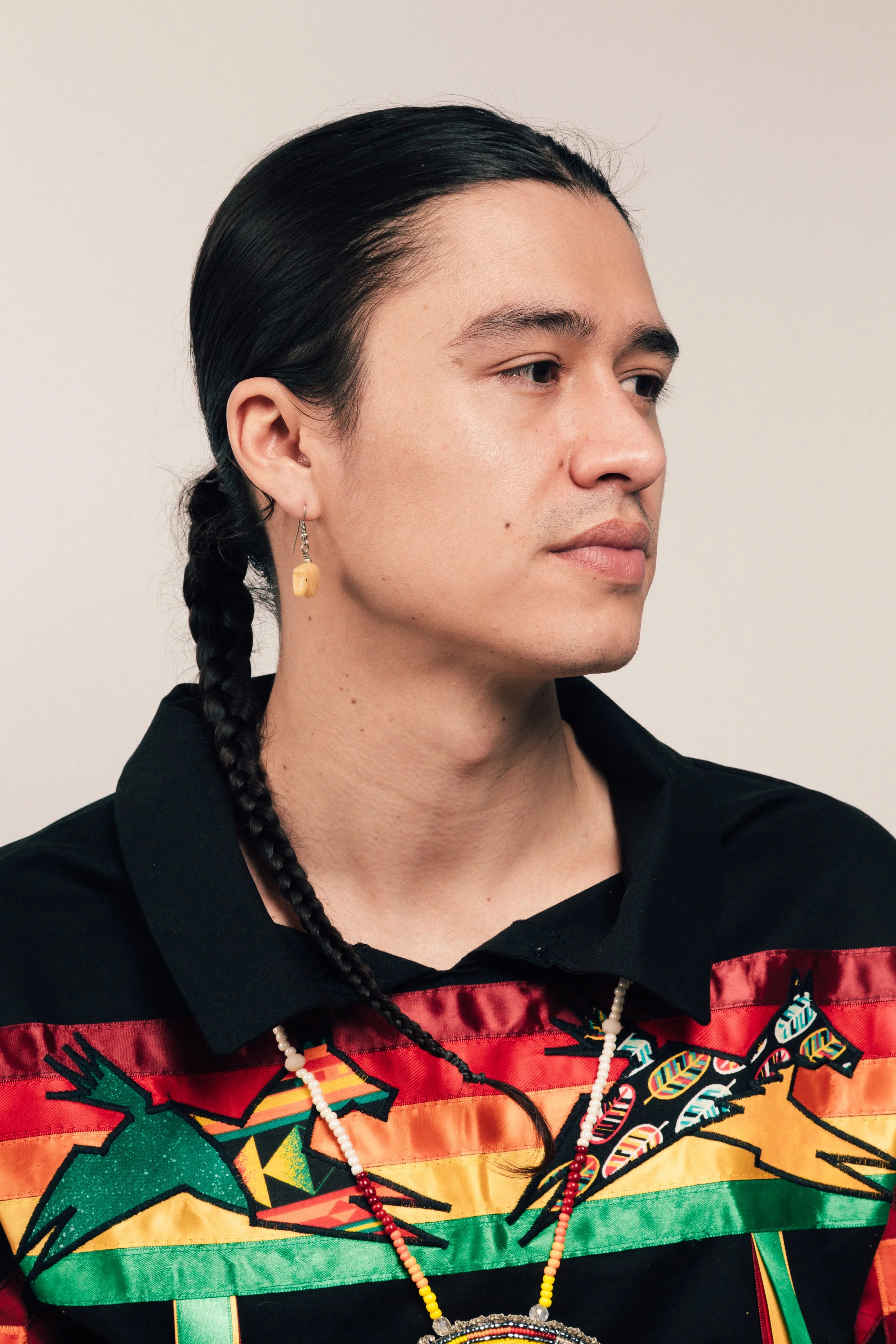 A person with long dark hair braided, wearing a colorful shirt with indigenous pattern, a beaded necklace, and an earring, against a plain background.