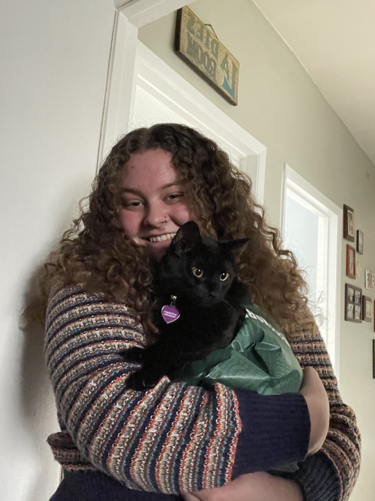 A woman with curly hair smiling while holding a black cat. The woman is standing indoors near a door with framed photos on the wall behind her.